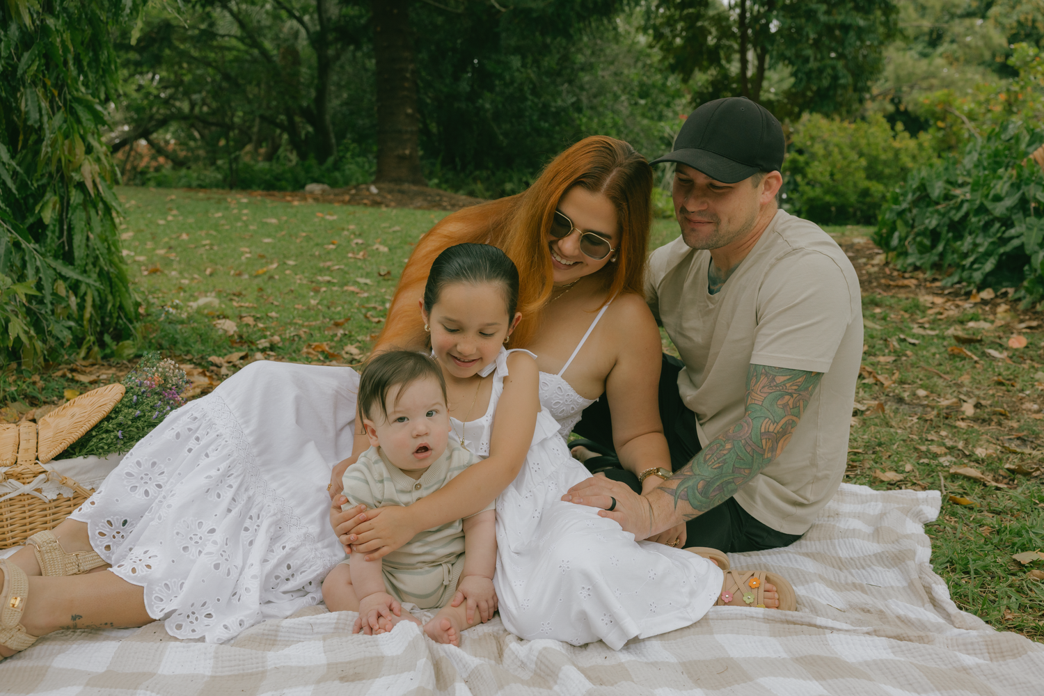 Family enjoying a picnic together on a white blanket under green trees on a sunny day.