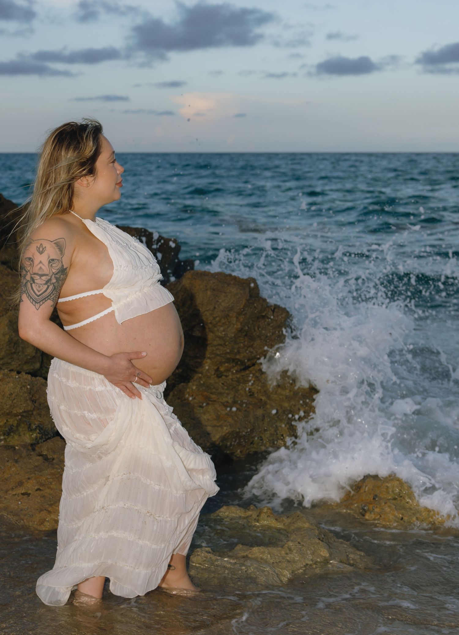 Pregnant woman in white dress stands on coastal rocks as waves crash against shore at sunset.