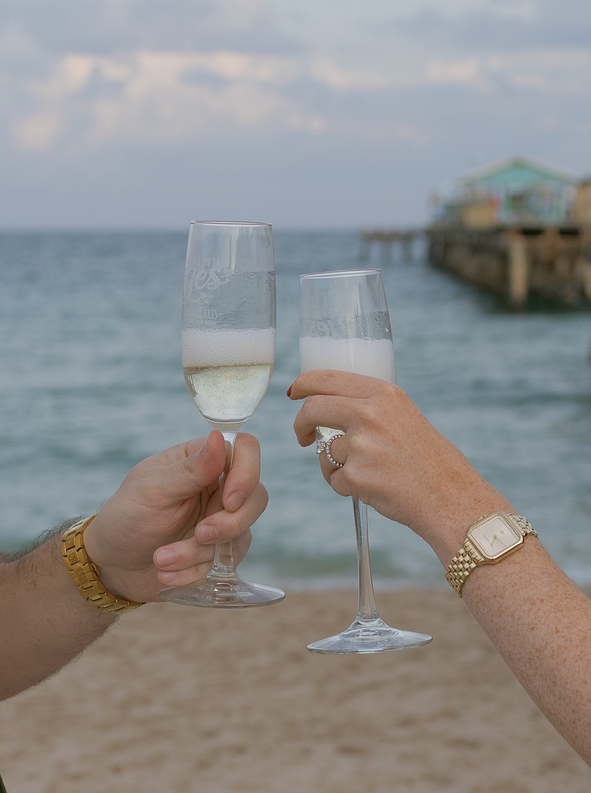 Two hands hold champagne glasses in a toast against a scenic beach and ocean backdrop during sunset.