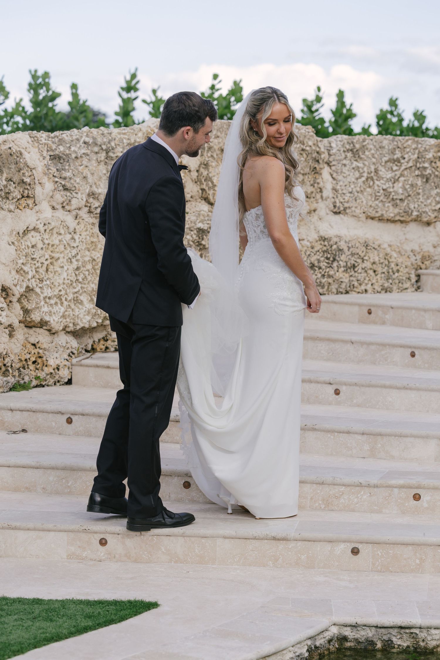 A couple in wedding attire walk down stone steps together in an outdoor garden setting.