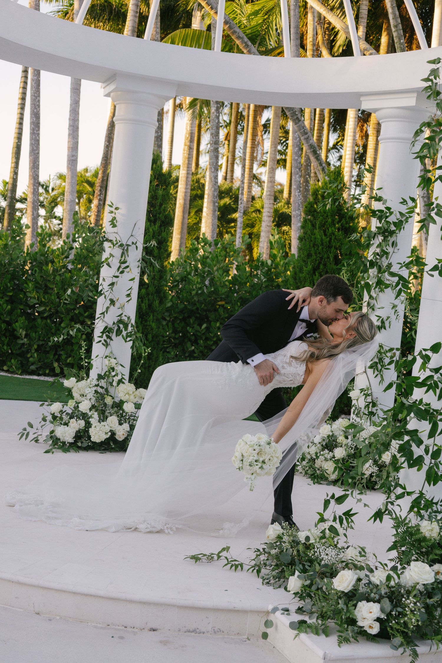 A romantic wedding scene with a couple embracing on a white lounge decorated with cascading greenery and palm trees.