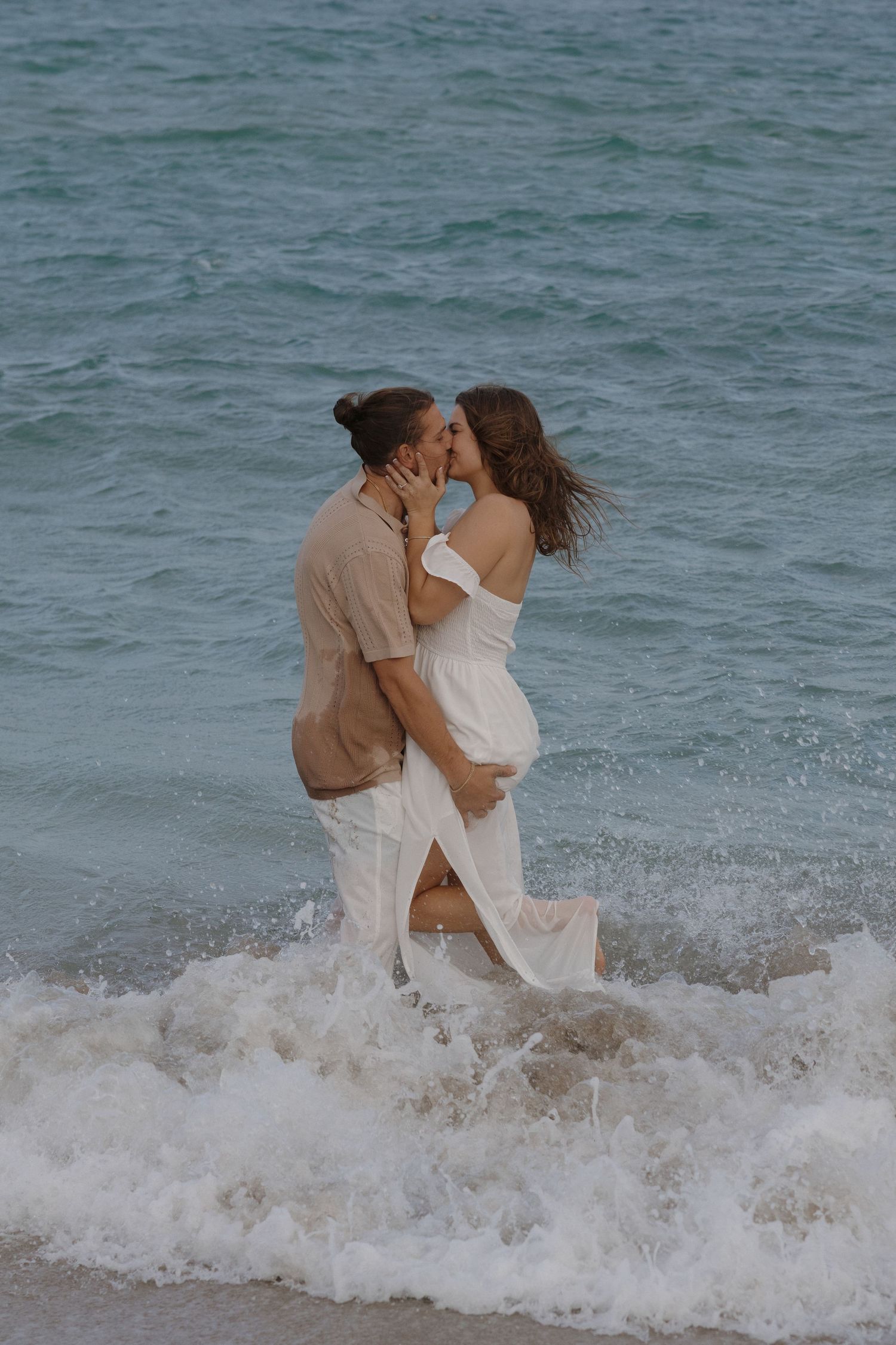 A couple embraces romantically on the beach as ocean waves splash around their feet at sunset.