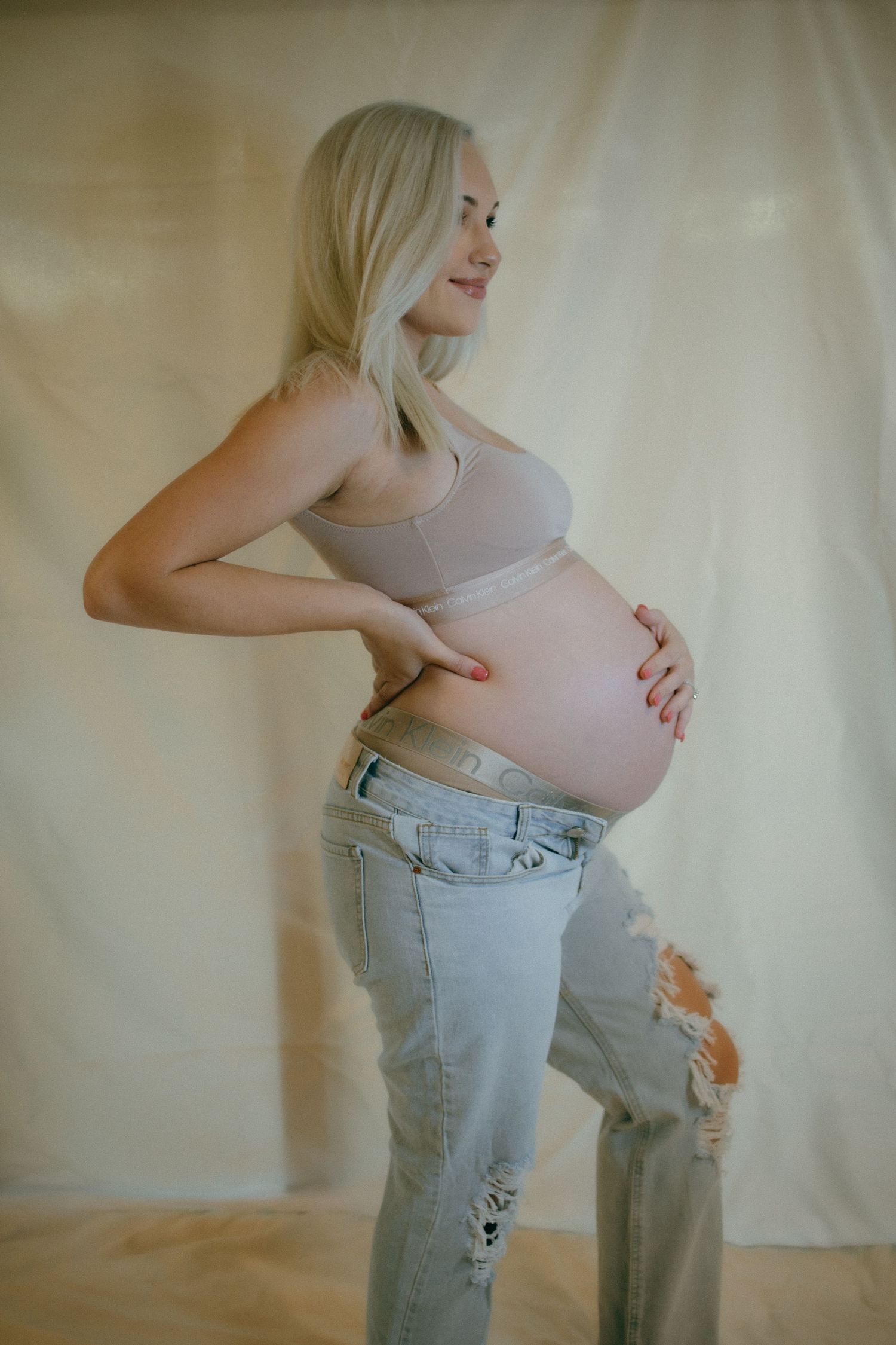 Side profile maternity photo with woman in casual attire against beige backdrop.
