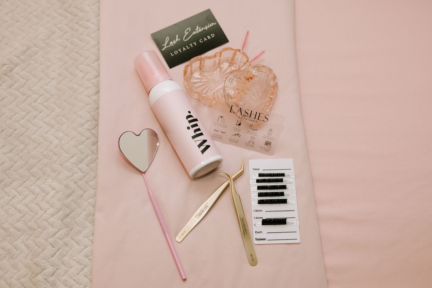 Flatlay of beauty products with gold pen and heart decoration on pink background.