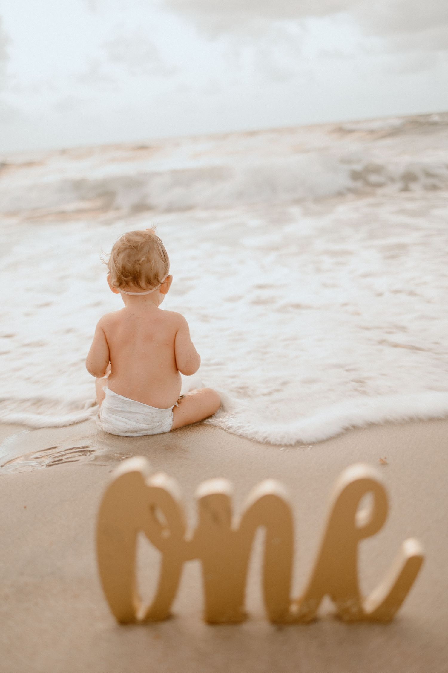 Baby sitting on sandy beach with 'one' text spelled out in wooden letters as ocean waves roll in behind.