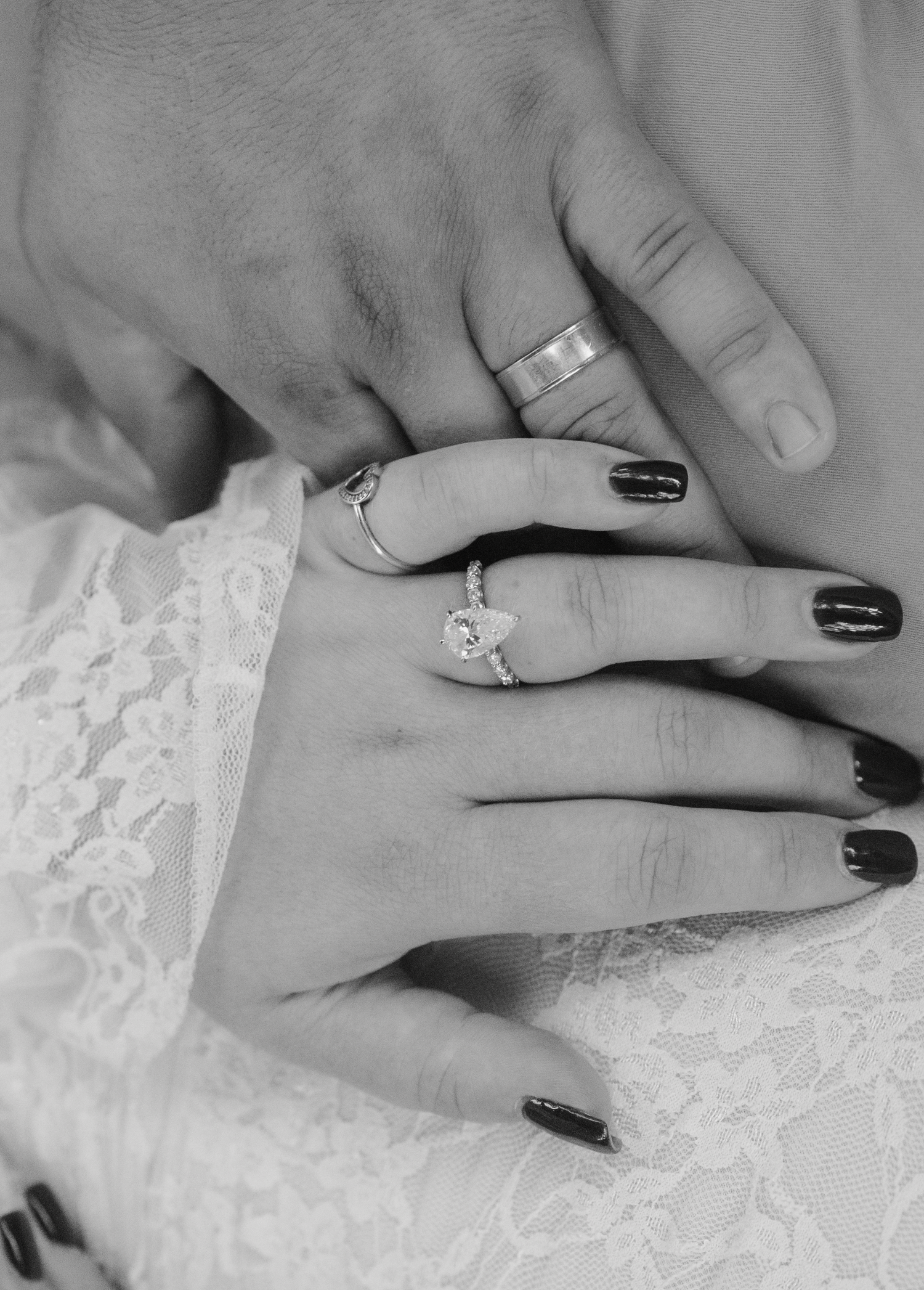 Black and white photo of hands wearing rings and dark nail polish resting on white lace fabric.