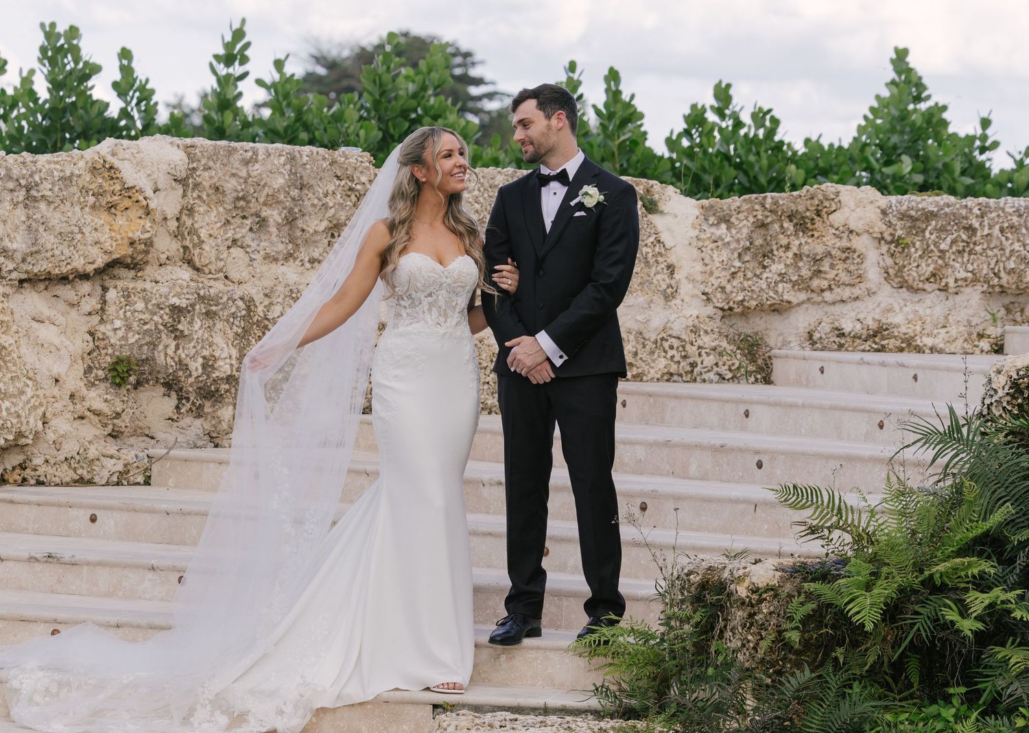 A couple shares a romantic moment in a garden setting with white stone steps and lush greenery in the background.