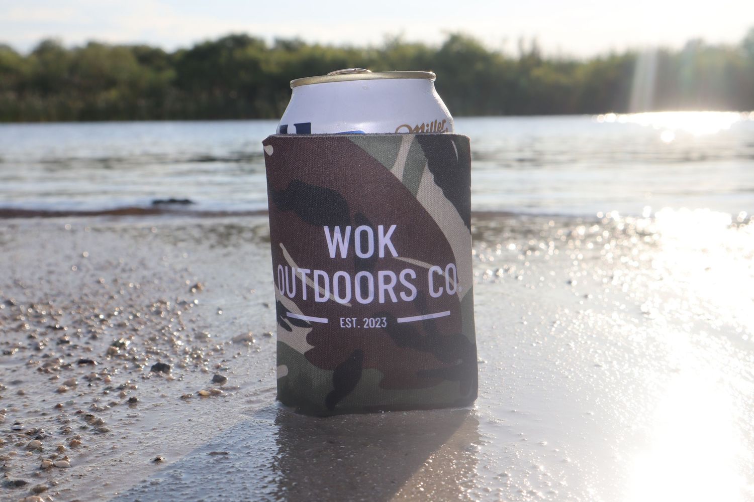 Camo koozie for WOK Outdoors Co. est. 2017 displayed on a sandy riverbank with water in the background.