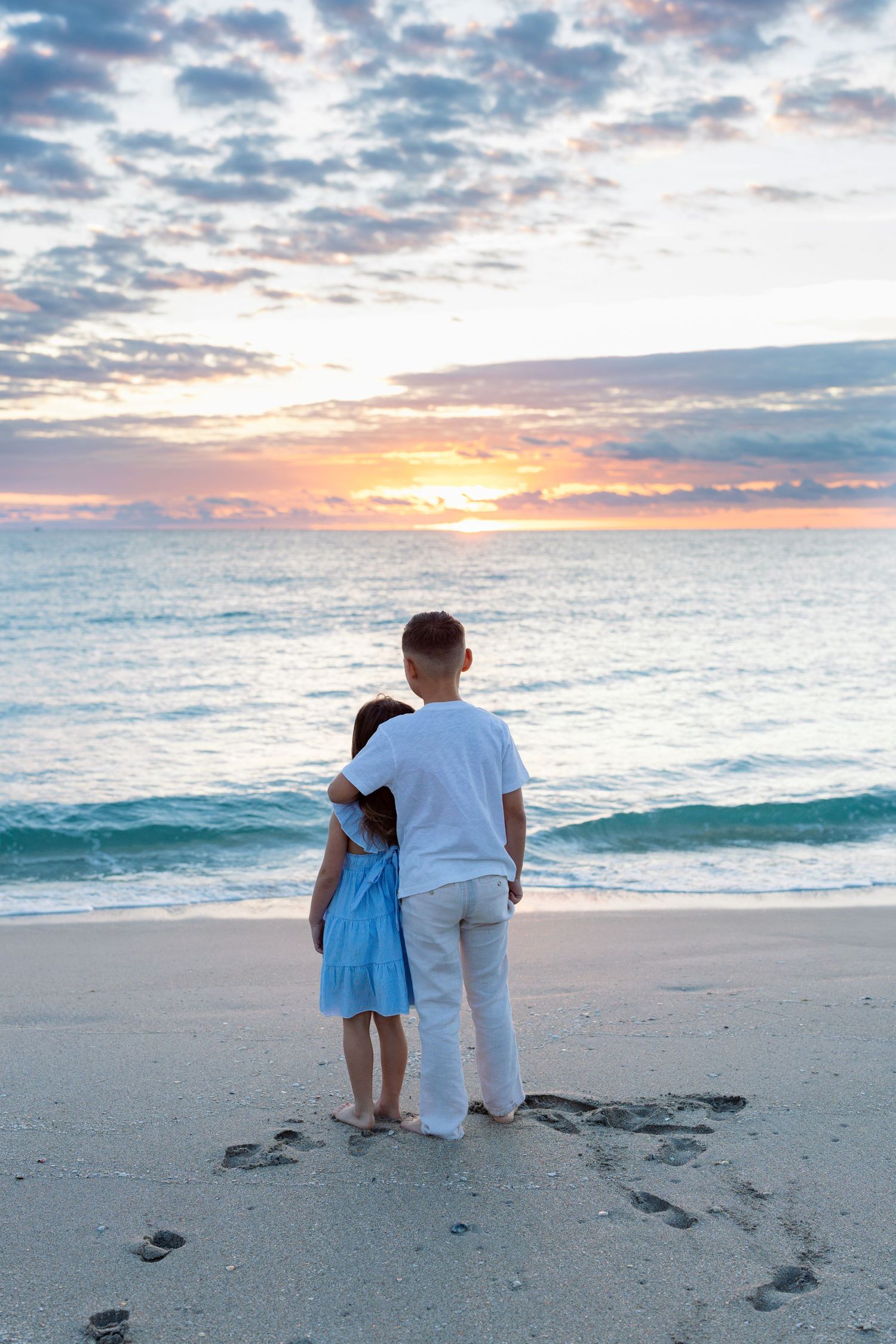 Two people sit together on a sandy beach watching a beautiful sunset over turquoise ocean waters.