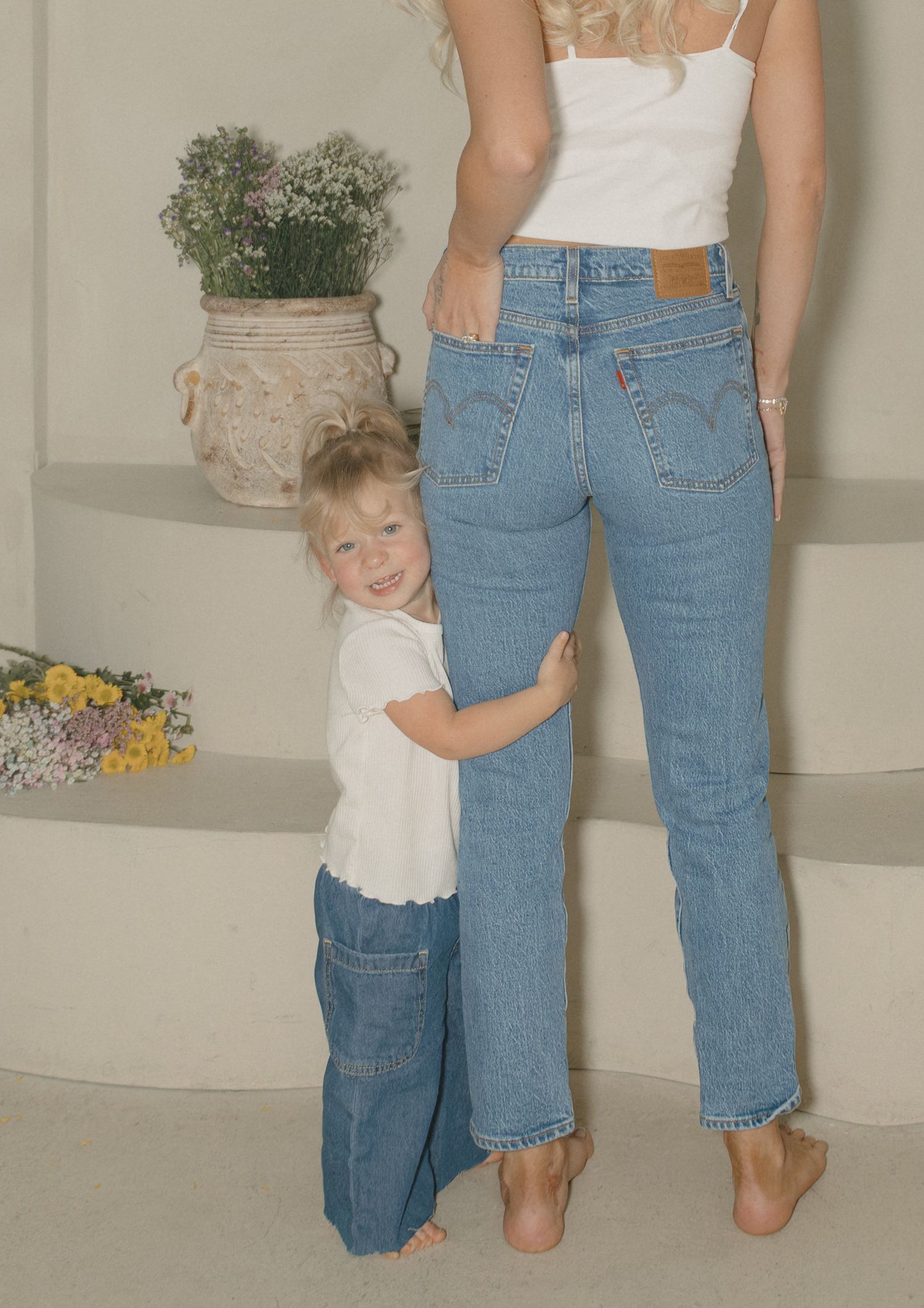 Young child hugging mother's legs, both wearing Levi's jeans, standing barefoot indoors near potted plants.