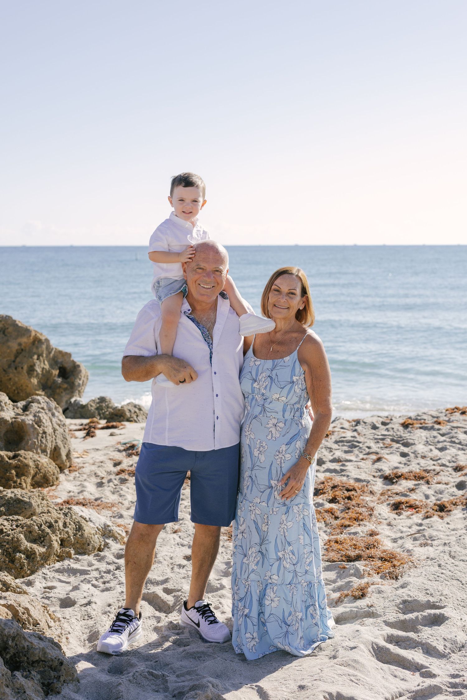 A family poses together on a sunny beach with rocky outcrops and ocean waves in the background.