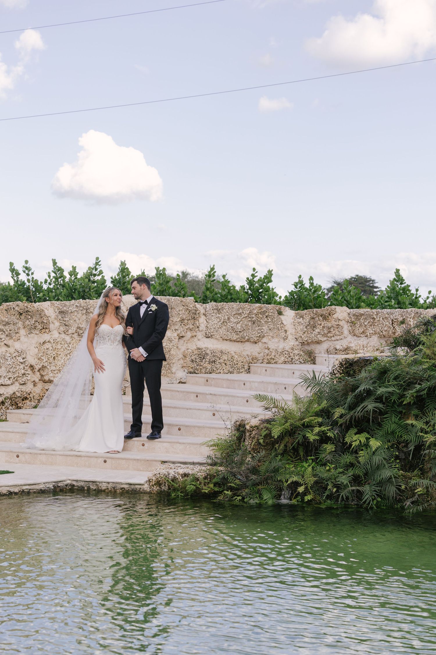 A couple in wedding attire poses together on stone steps overlooking a serene pond surrounded by greenery.