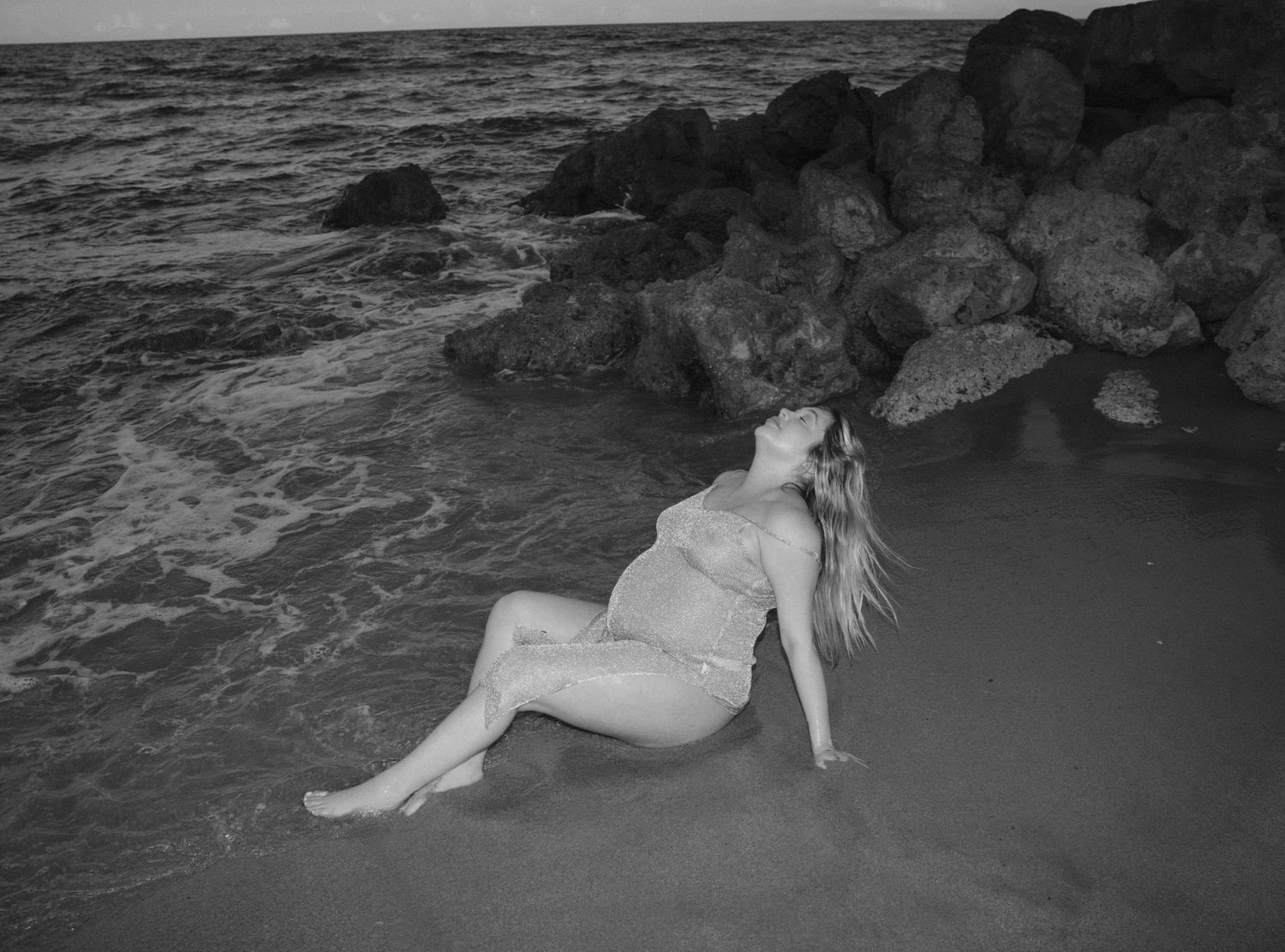 Artistic black and white maternity portrait on beach with dramatic waves and rocks.