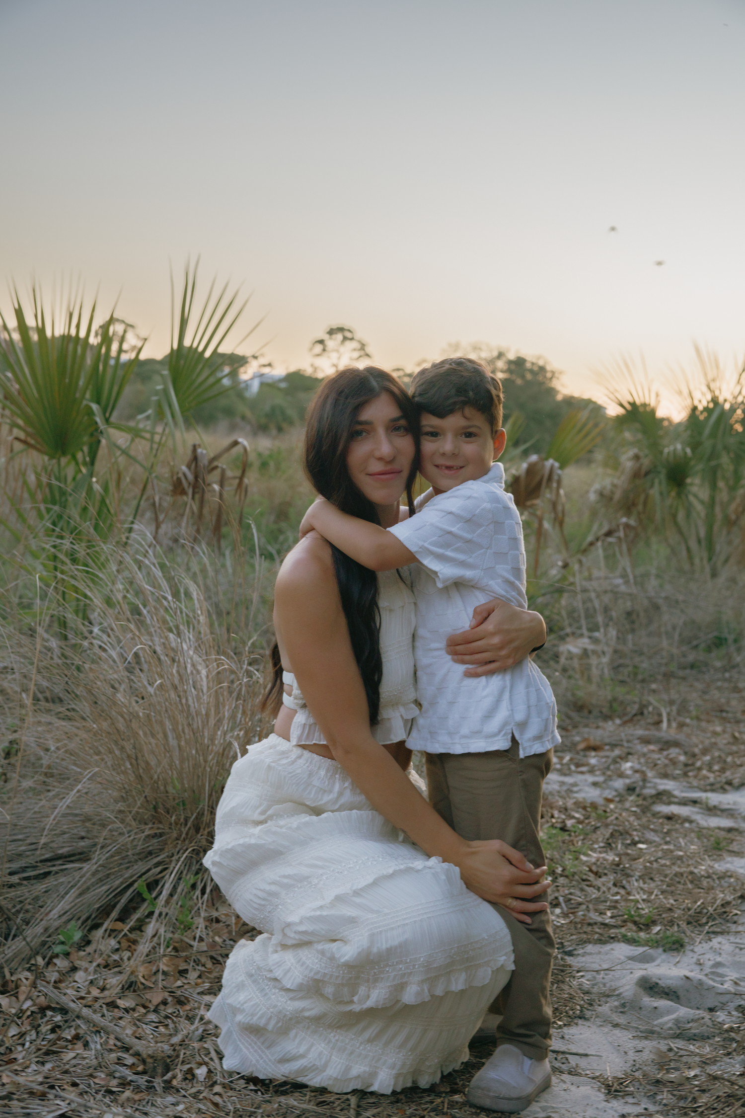 A heartwarming outdoor portrait of a mother and child embracing among tall grasses at sunset.