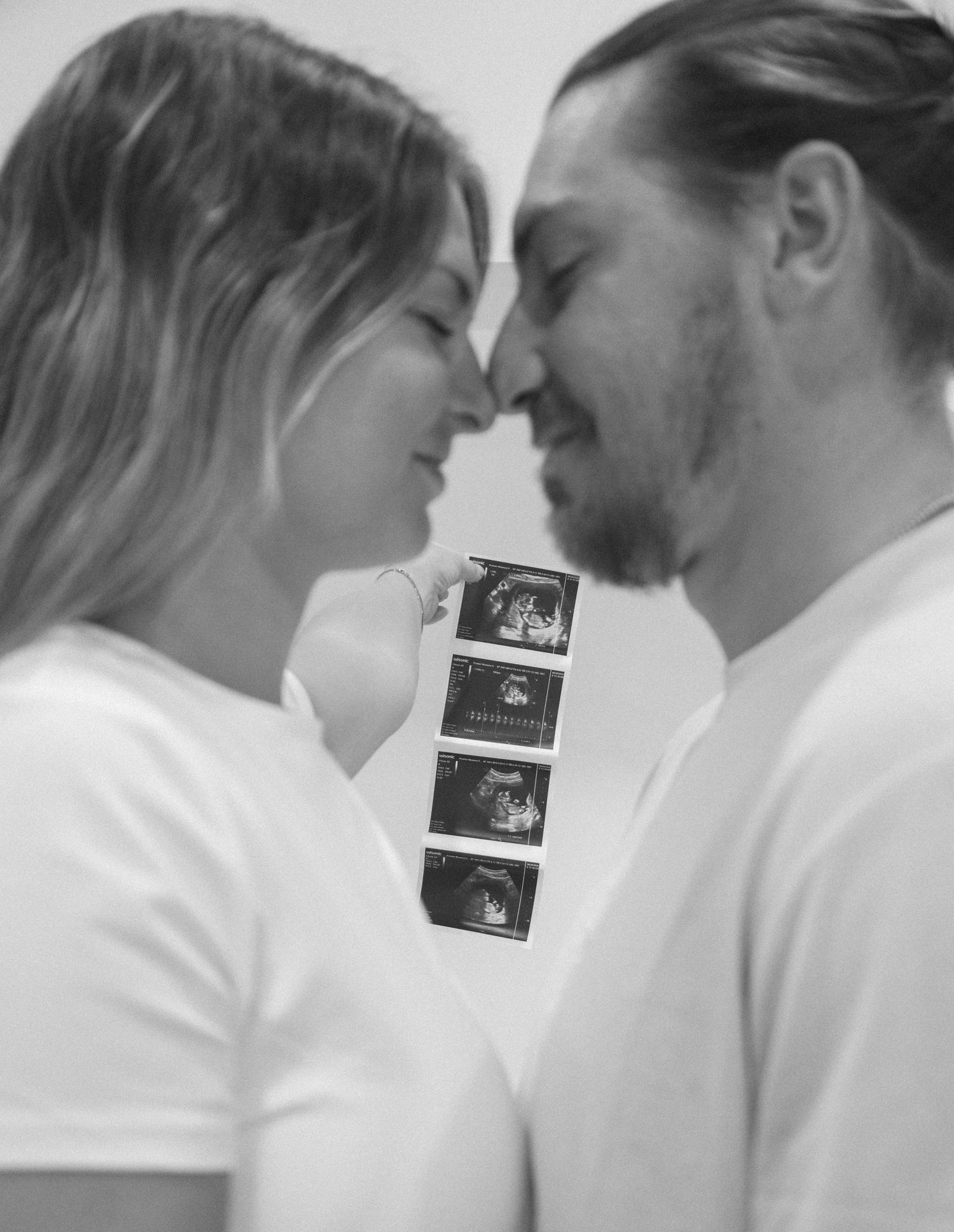 Black and white photo of a couple touching noses, smiling while holding ultrasound images, announcing their pregnancy.