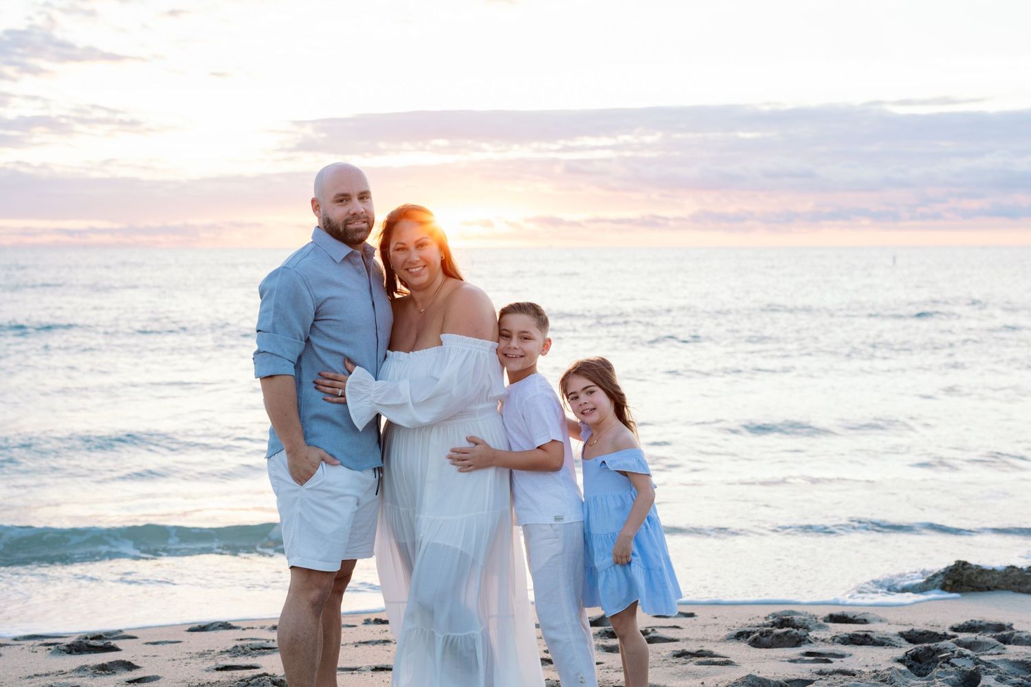 A group in white and blue outfits poses together on a beach at sunset for family portraits.