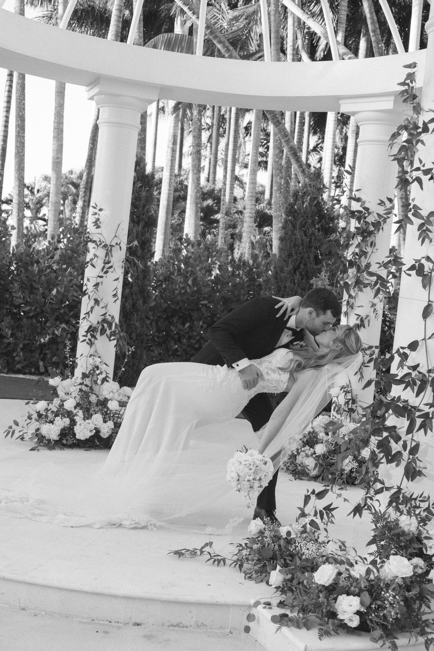 A romantic dip pose captured during a wedding photoshoot surrounded by tropical palm trees and white floral arrangements.