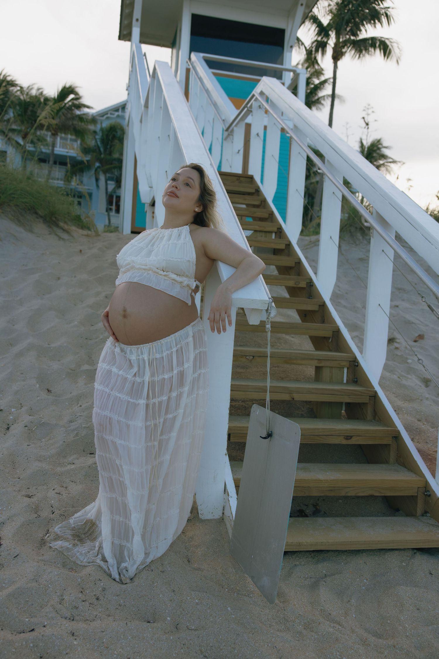 A figure in a white flowing dress poses artistically on sandy beach steps during a photo shoot.