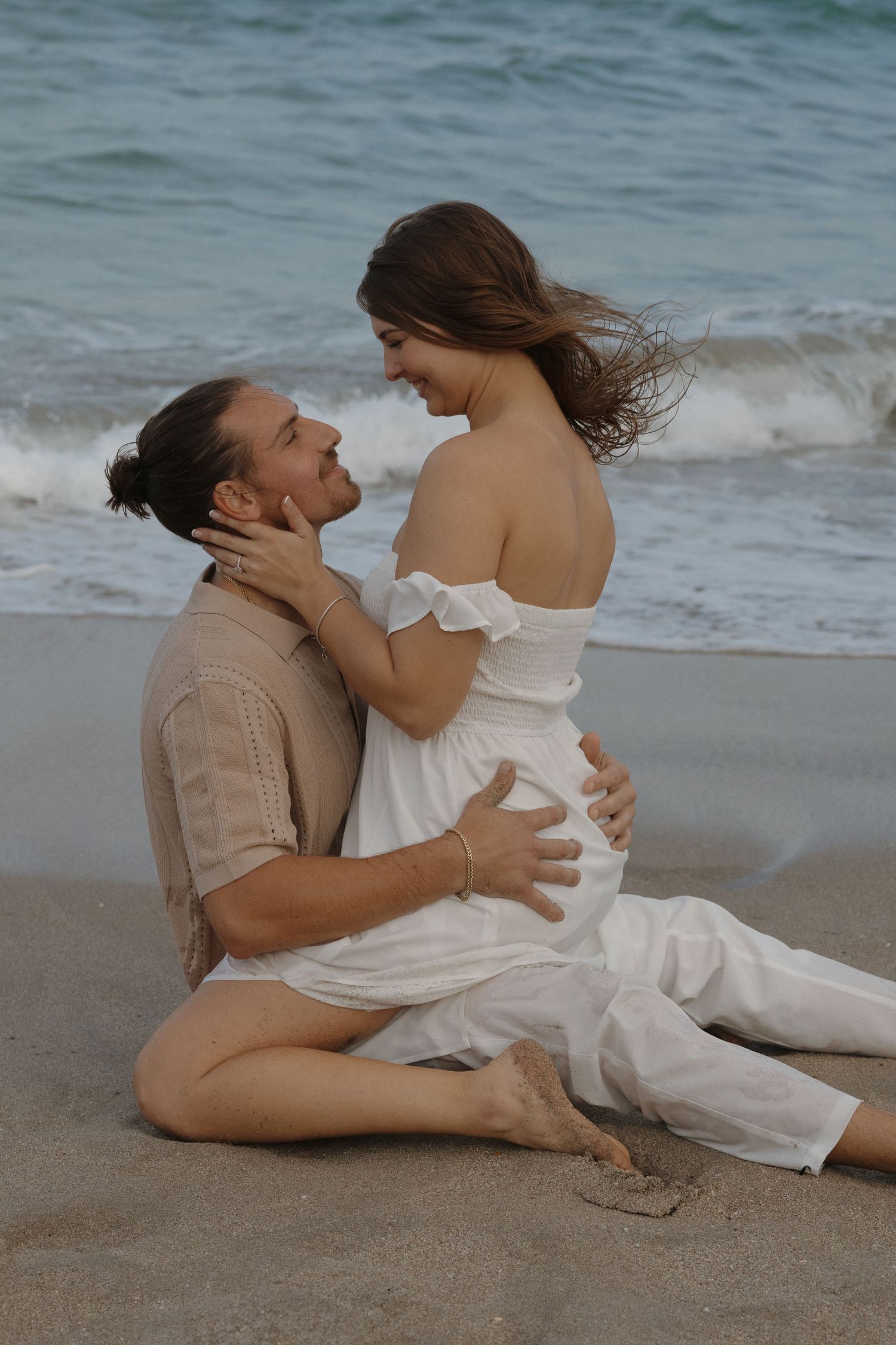 Romantic couple shares intimate moment sitting on beach with crashing waves in background at sunset.