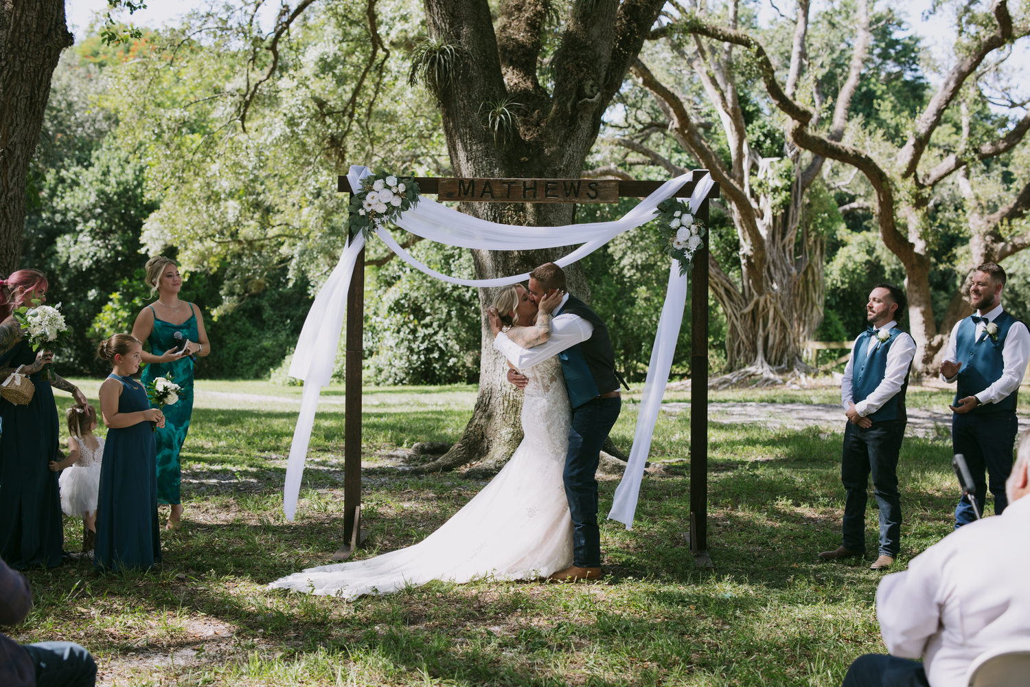 An outdoor wedding ceremony under a simple white fabric arch amongst towering oak trees on a sunny day.