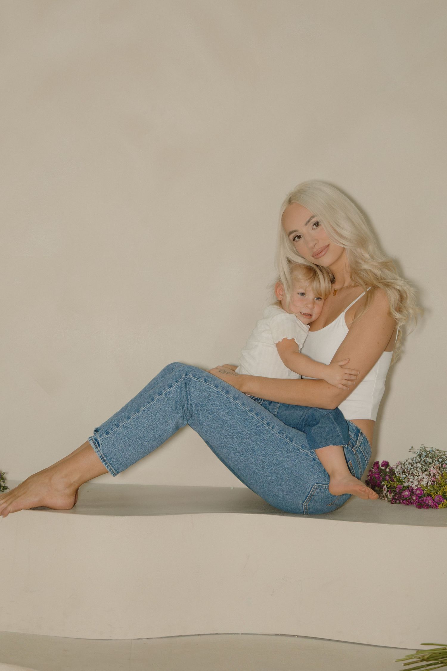 Blonde woman in white top and jeans sits barefoot holding a young child against a neutral background.
