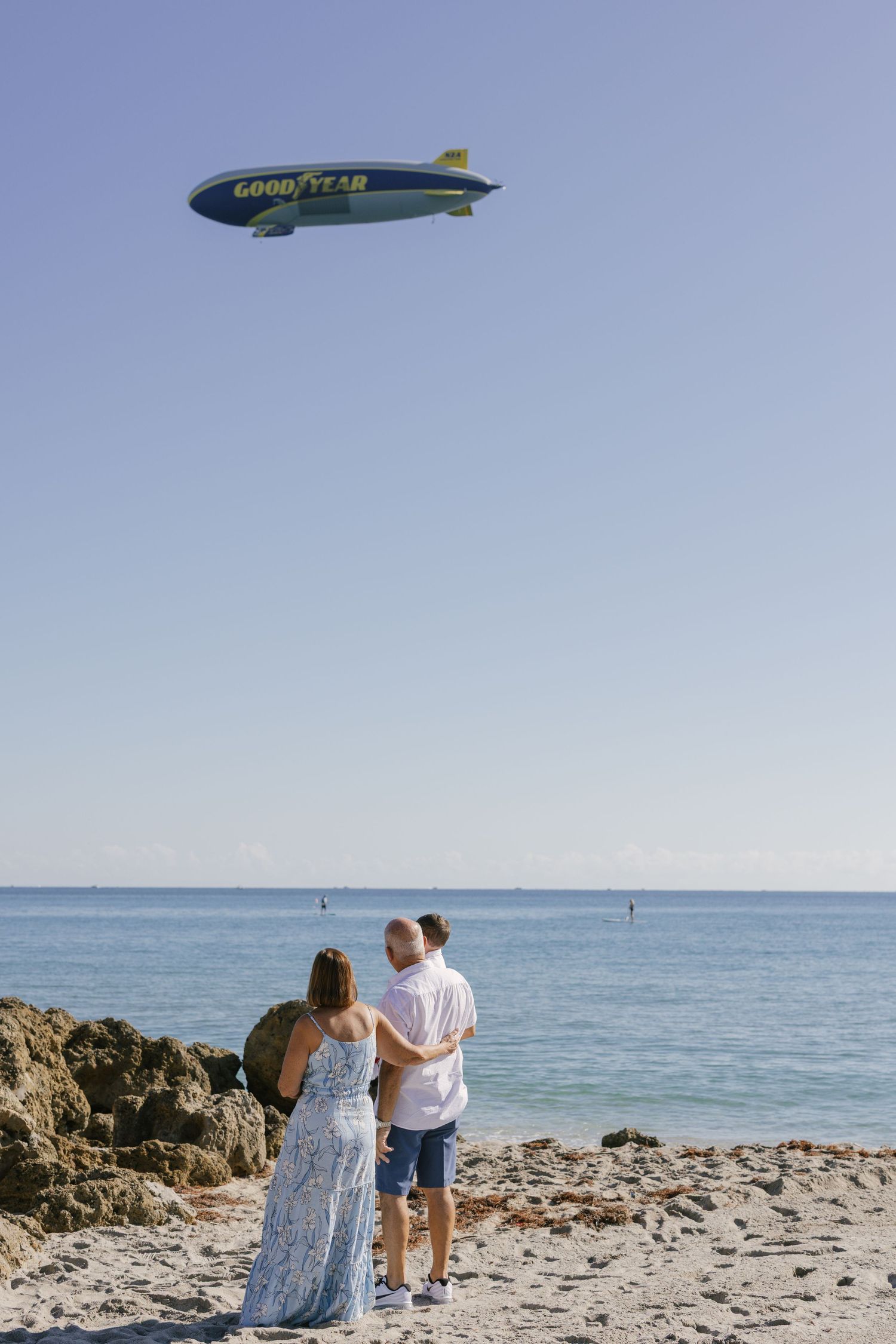 A couple walks along a rocky beach with a Goodyear blimp visible in the blue sky above the ocean.