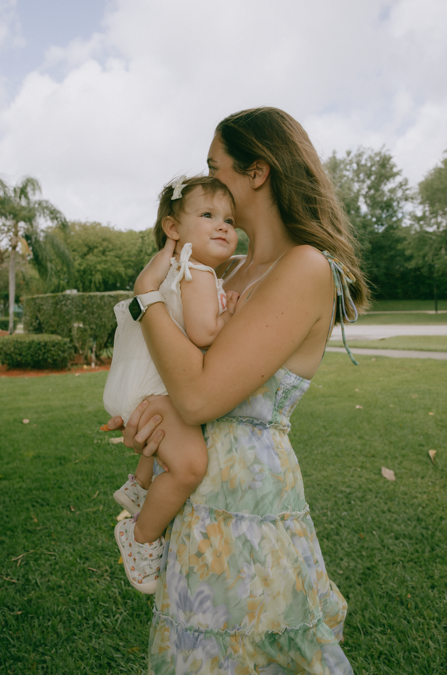 A tender moment captured outdoors in a garden setting with a floral dress and white outfit against lush green grass.