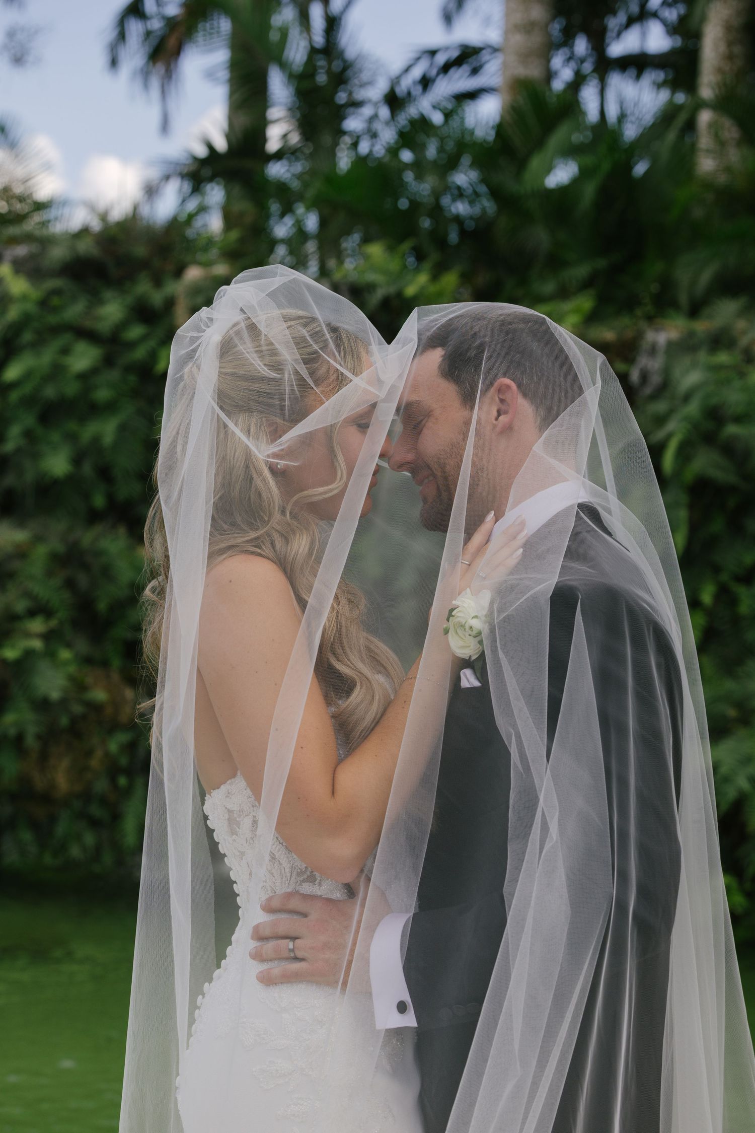 A romantic wedding couple shares a tender kiss under a flowing white veil in a lush garden setting.