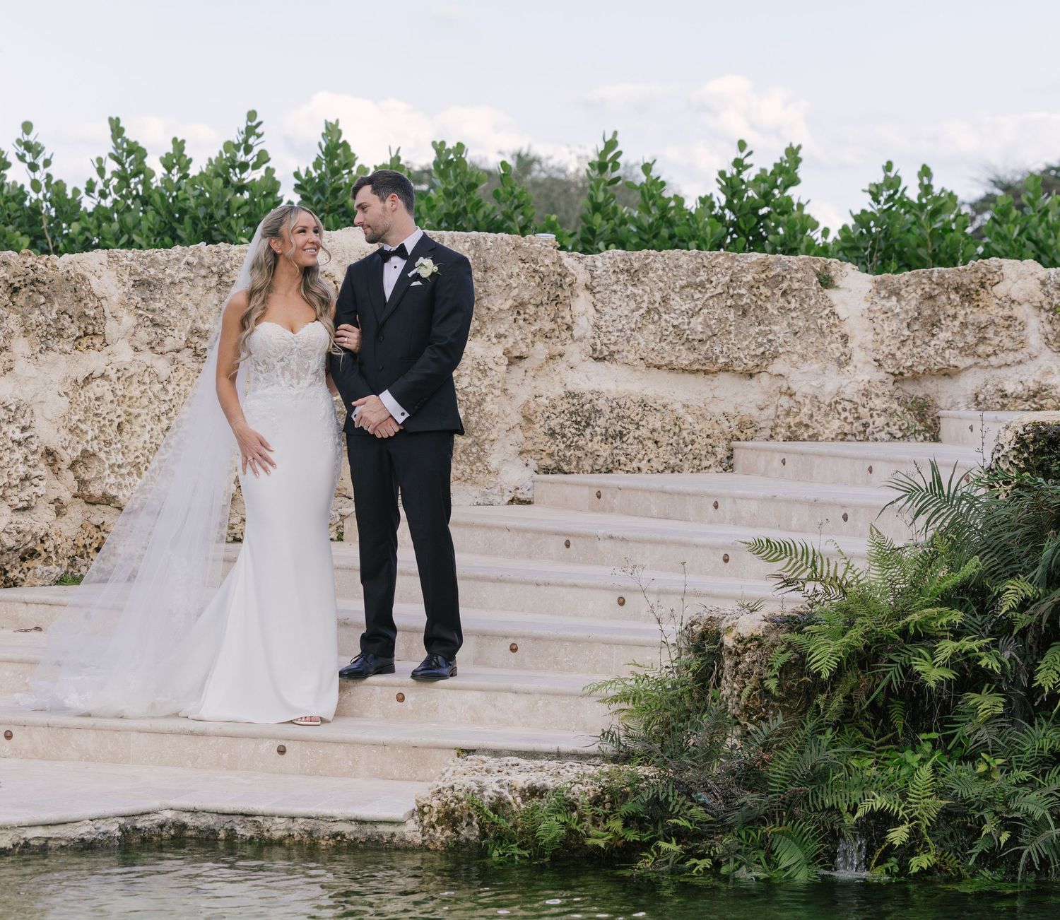 A romantic outdoor wedding couple poses on stone steps surrounded by lush greenery and gardens.