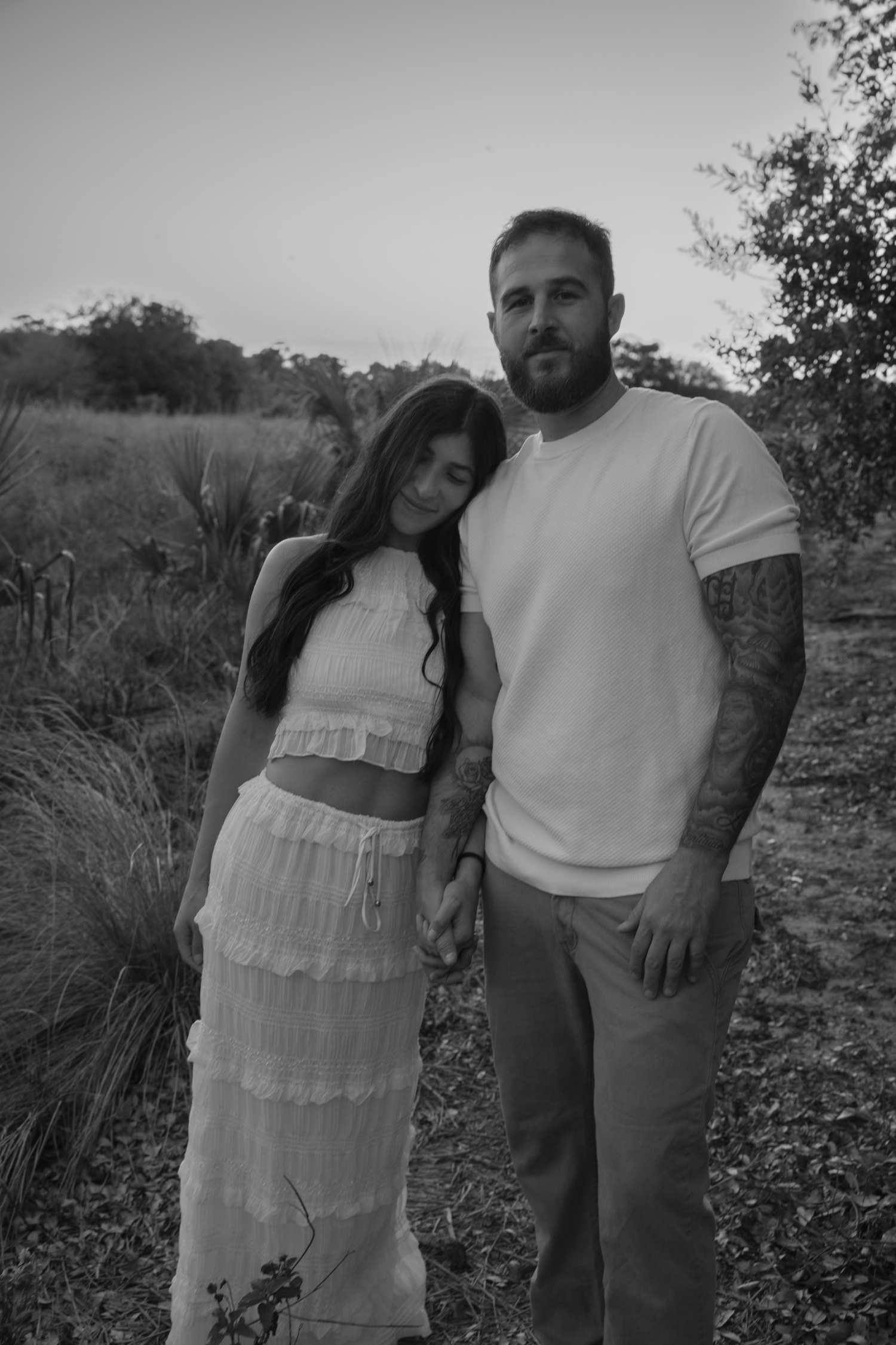 A couple poses together outdoors at sunset, with the woman wearing a white tiered dress and the man in a white t-shirt.