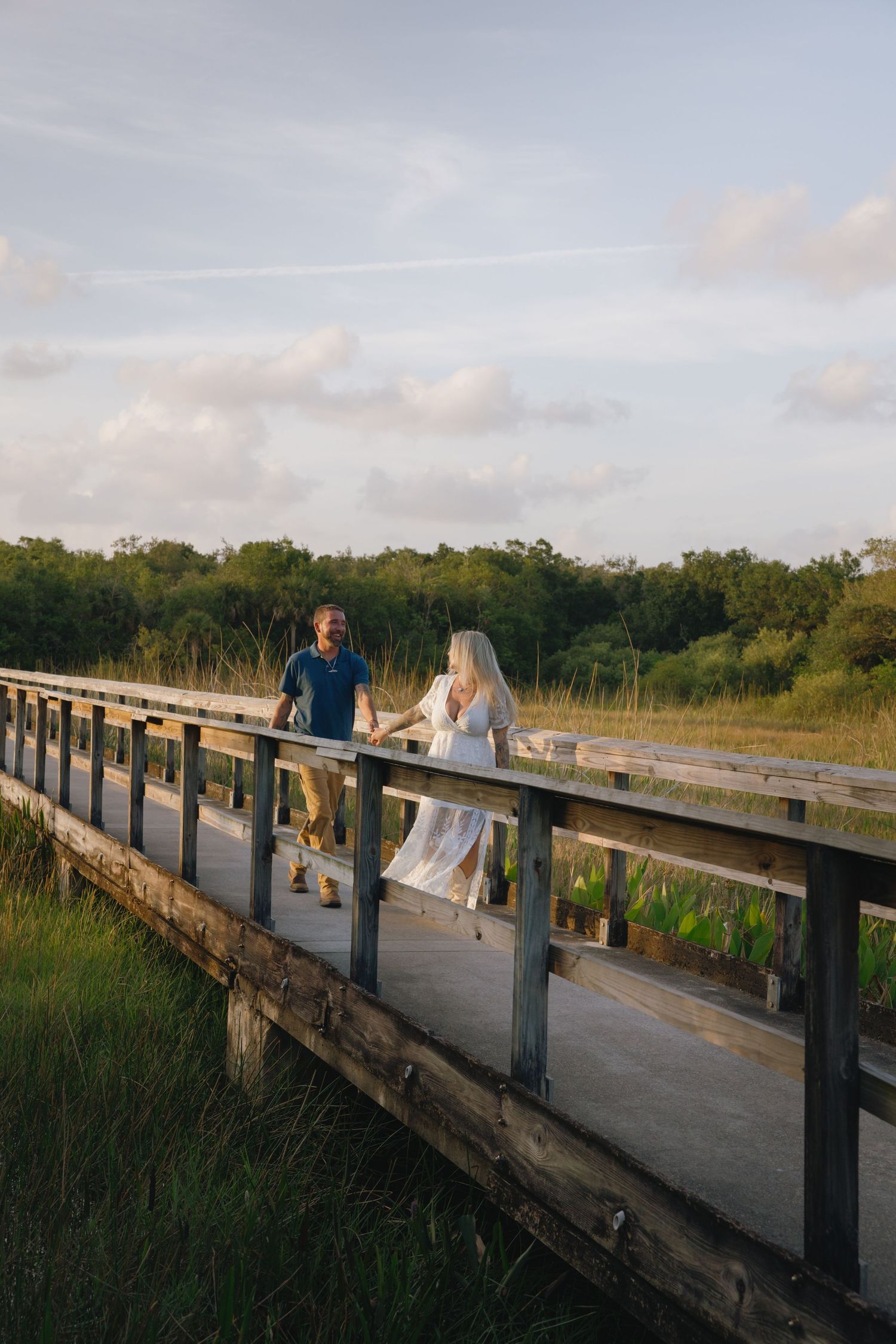 A couple walks together along a wooden boardwalk through natural marshland at sunset.