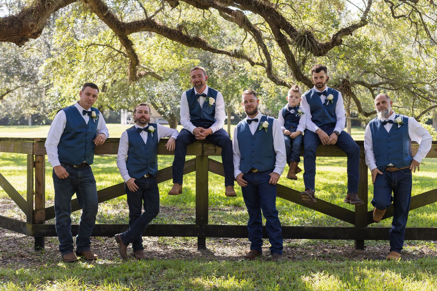 Wedding party in navy blue vests poses casually on a rustic wooden fence under large oak trees.