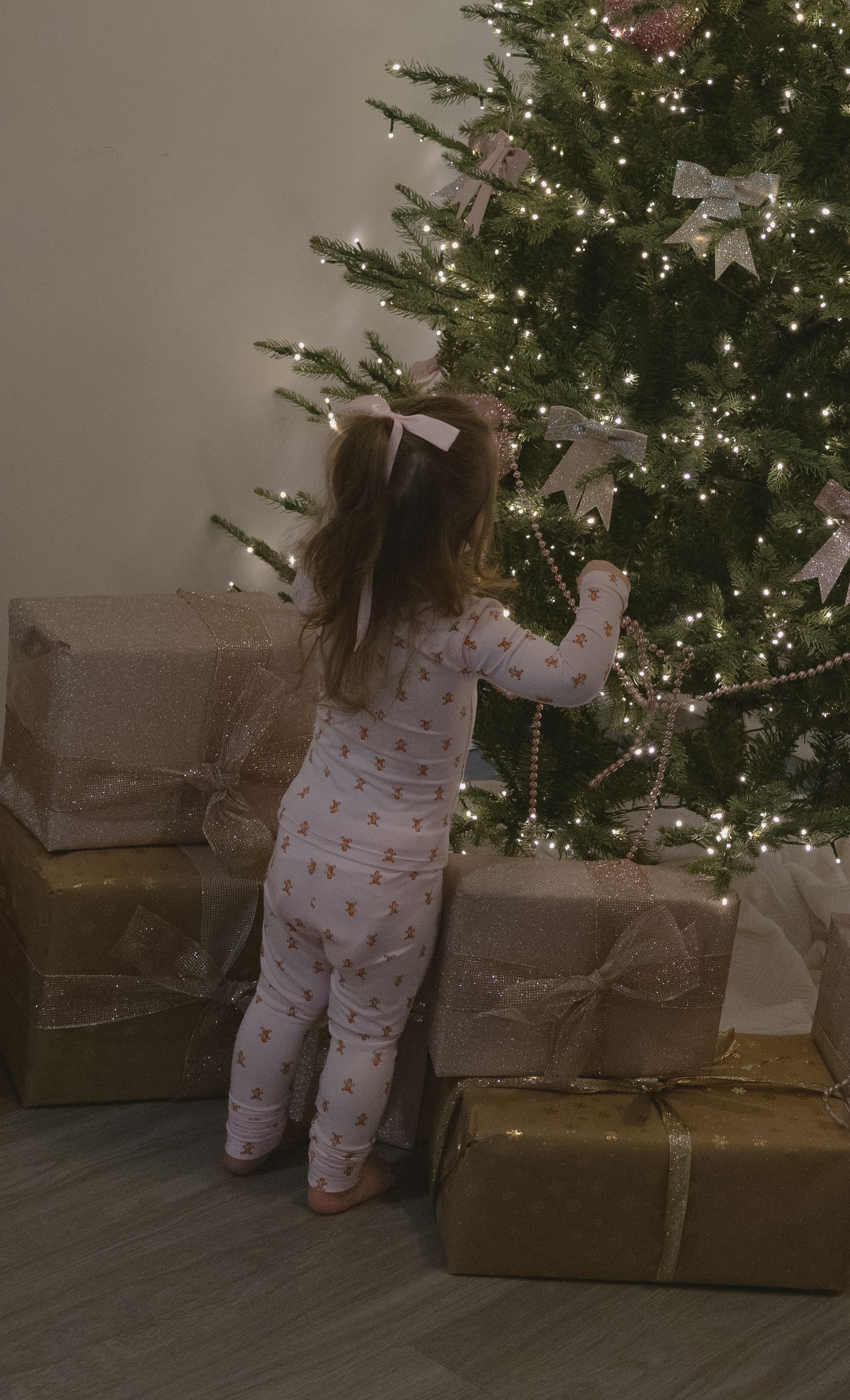 A small child in festive pajamas reaches up to touch a lit Christmas tree next to wrapped presents on the floor.