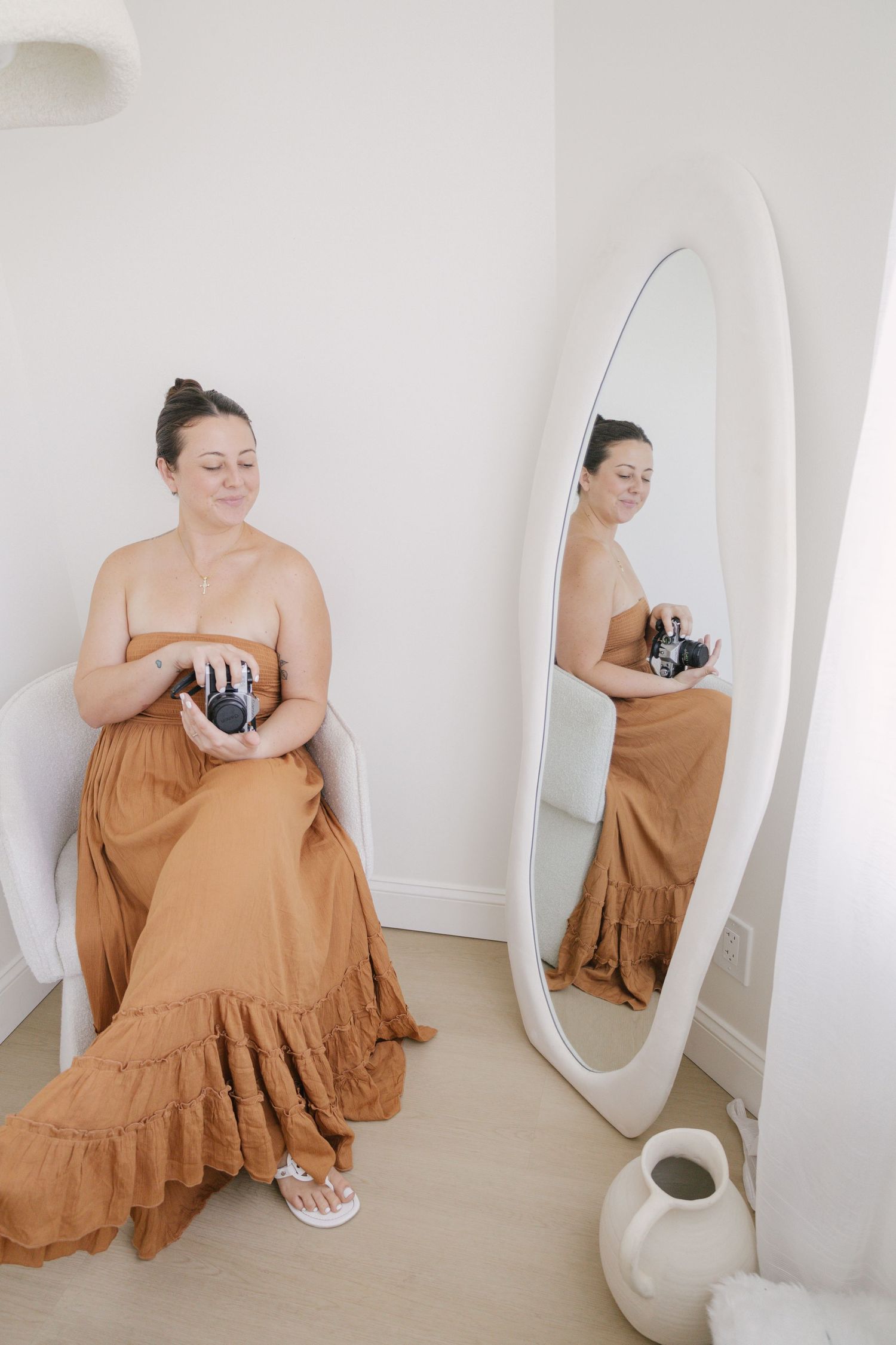 Woman in rust maxi dress holds camera beside oval mirror in bright white room.