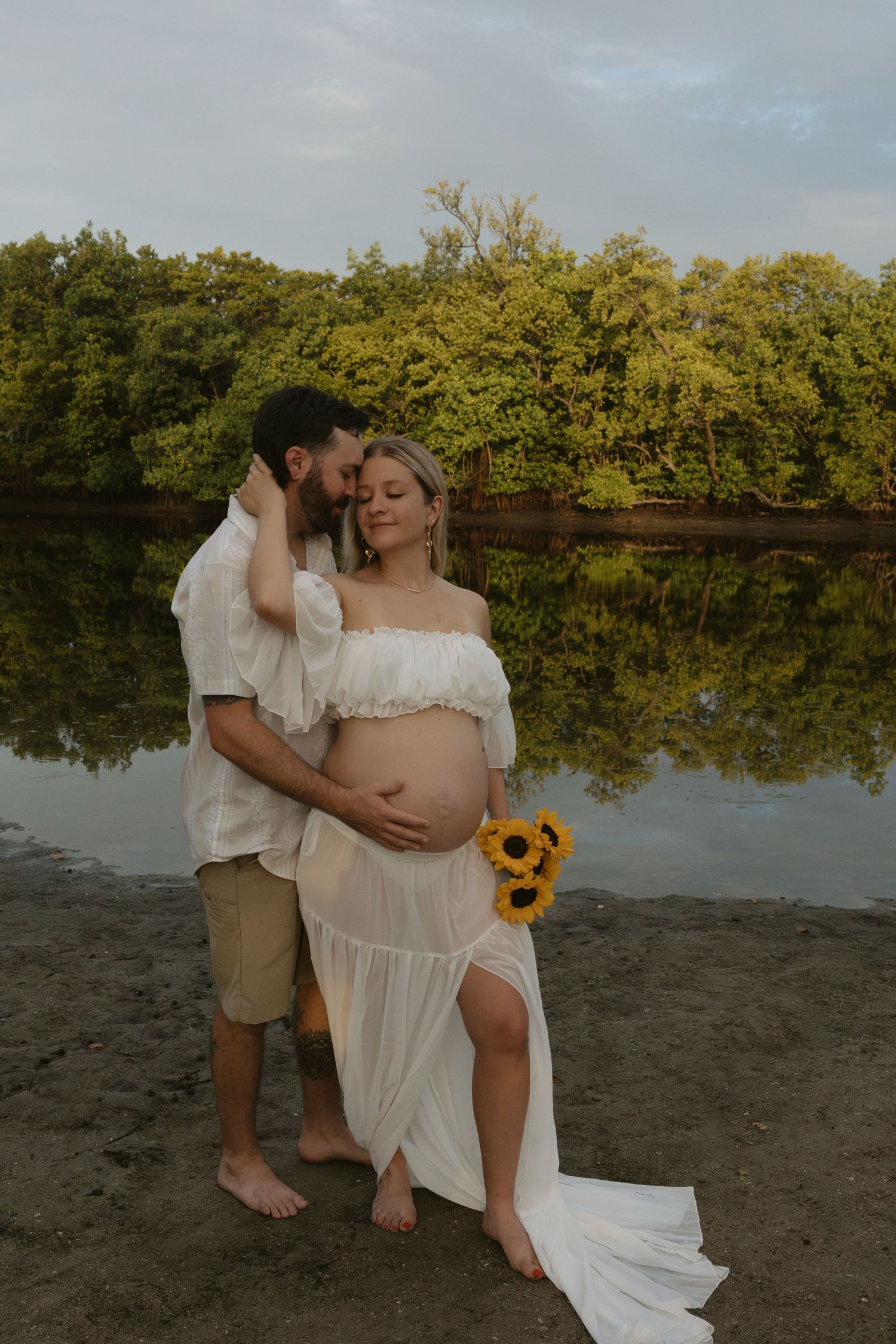 Romantic maternity photo session at sunset near a peaceful lake with lush greenery and reflective waters.