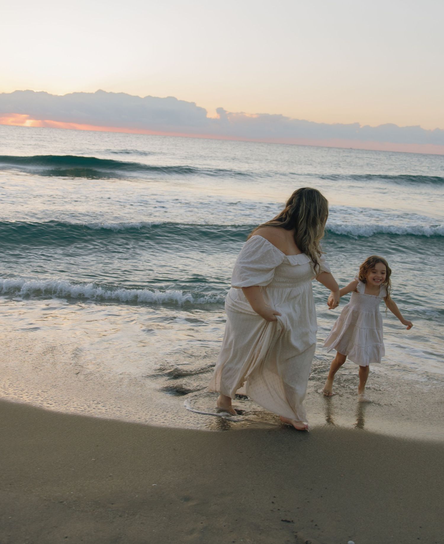 Two people in white dresses walk along the shoreline as waves crash at sunset.