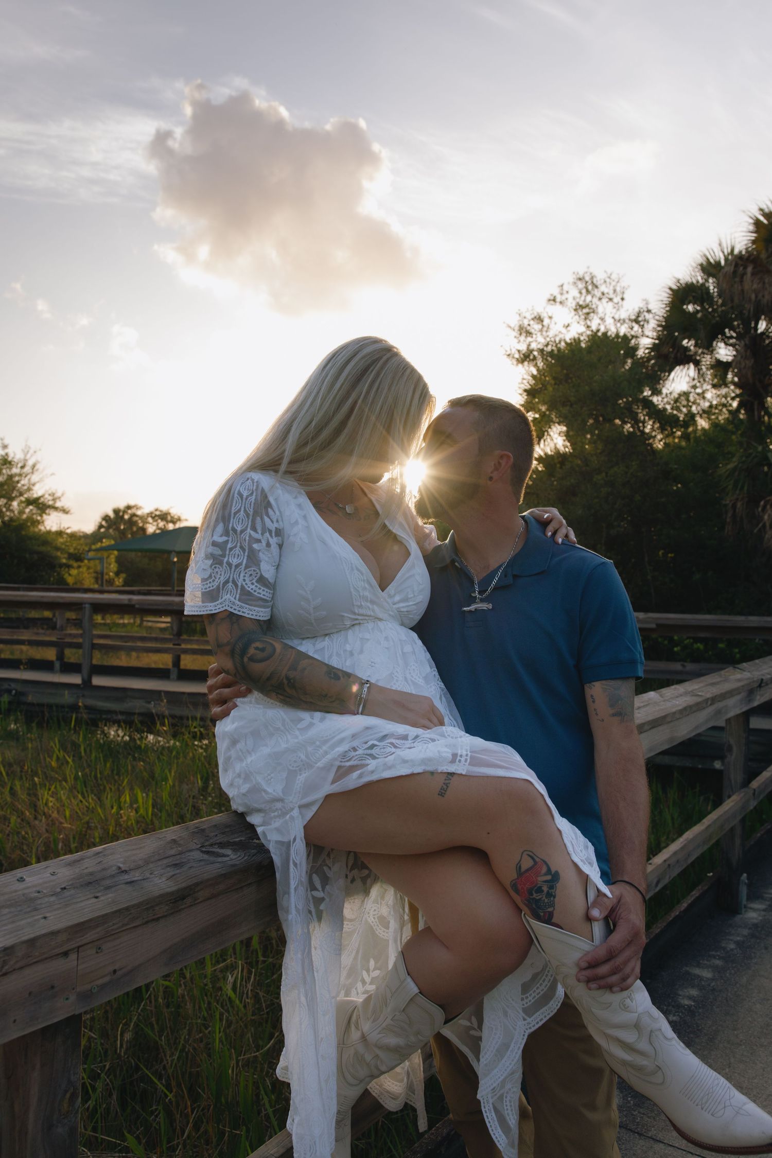 A romantic couple sits together on a wooden boardwalk at sunset with sun rays peeking through clouds.