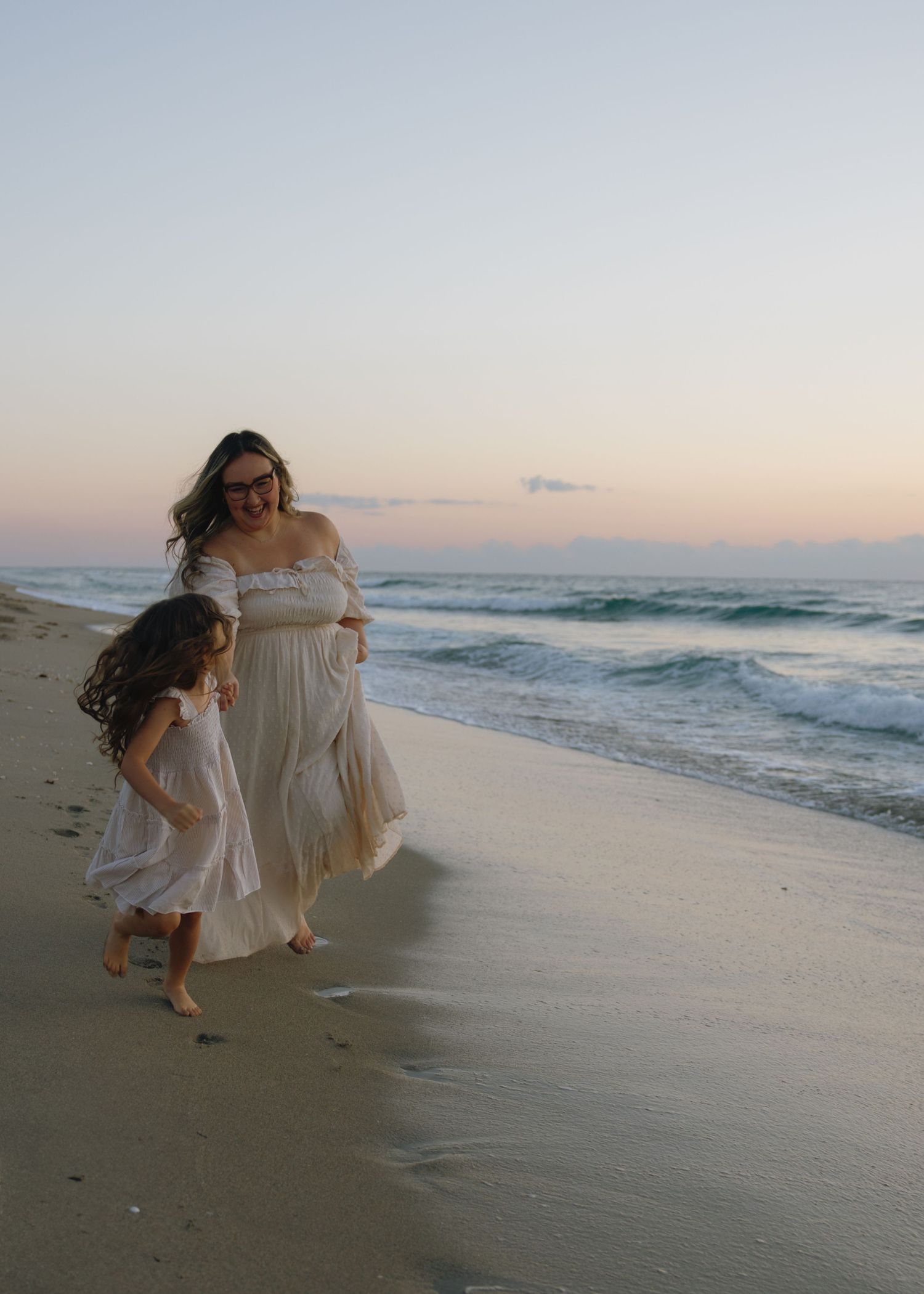Mother and daughter walking along beach at sunset in flowing dresses.