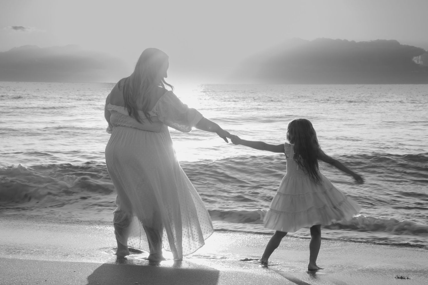 Two figures in white dresses dance together on a beach at sunset creating a dreamy black and white silhouette.