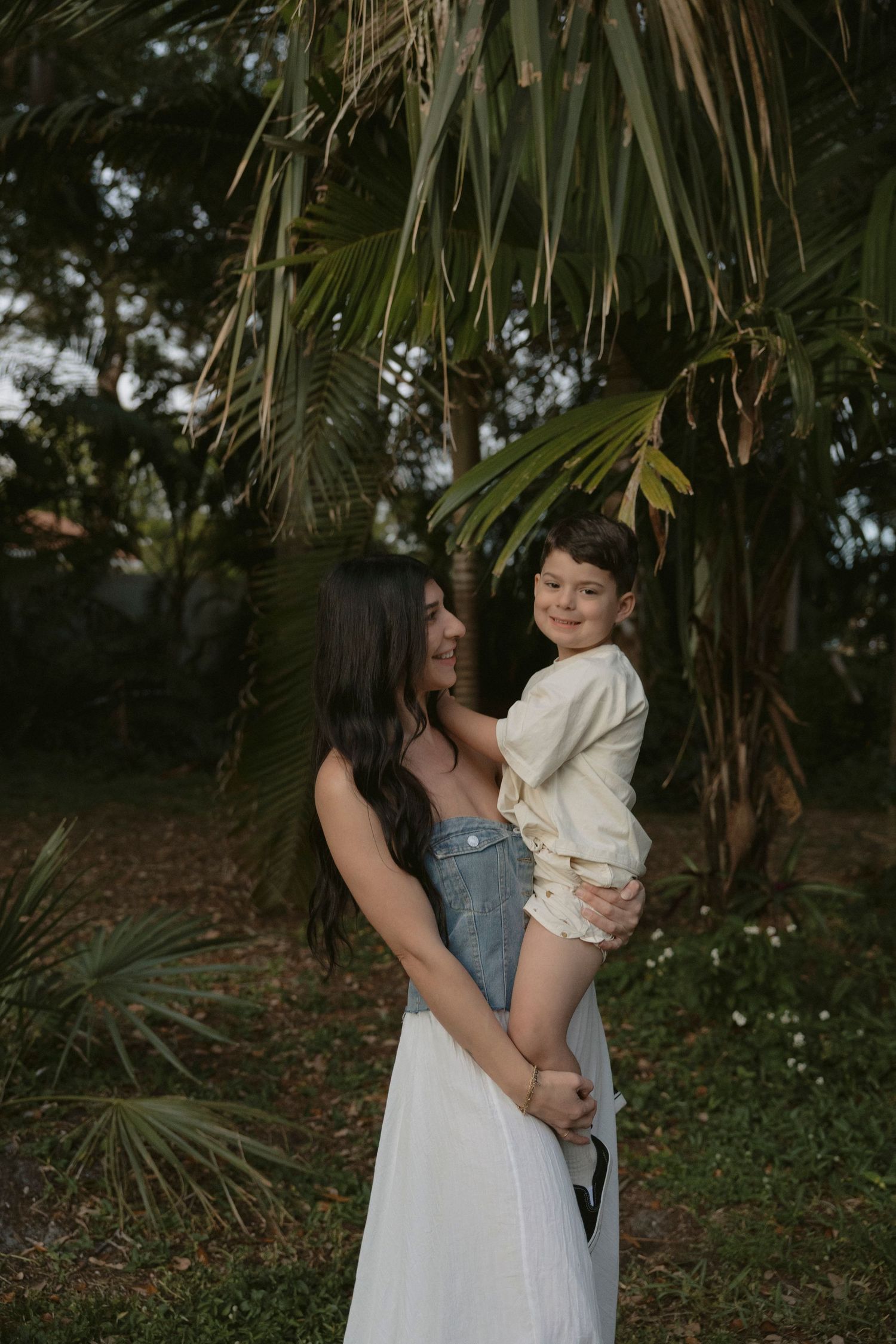 A tender embrace between two people in a lush tropical garden setting with palm fronds in the background.