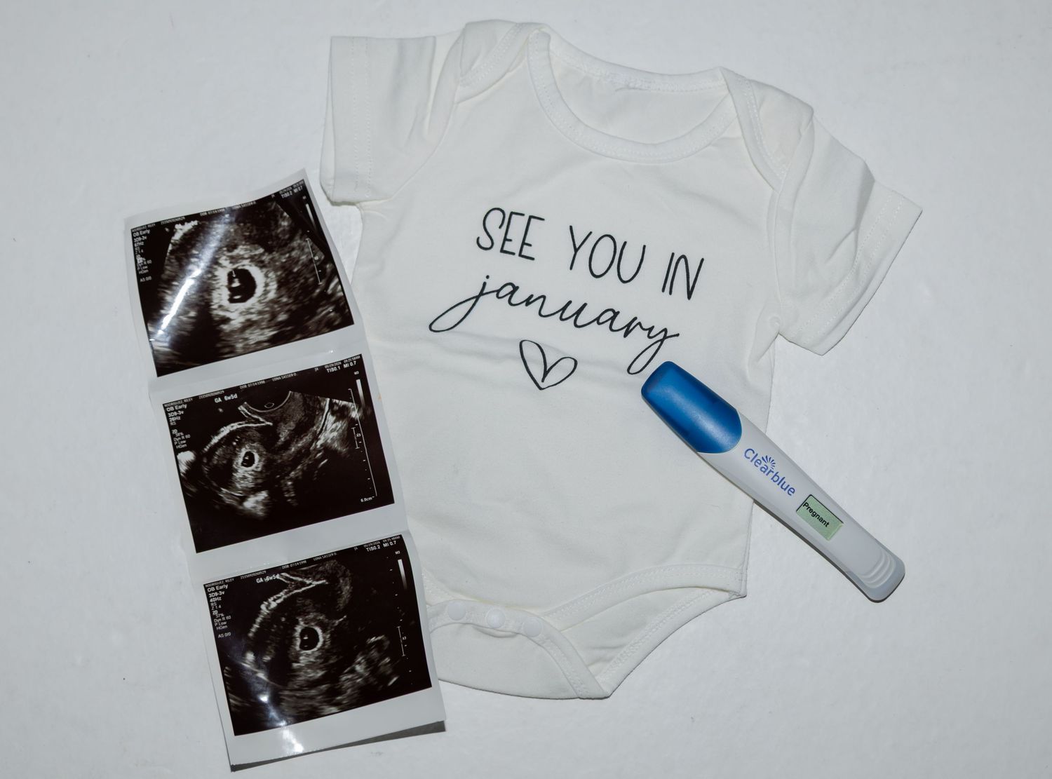 Baby announcement onesie saying See You In January with ultrasound photos and positive pregnancy test on white background.
