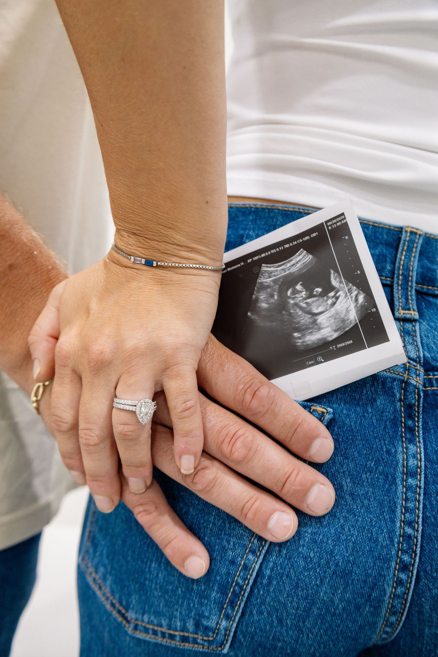 Couple holding hands with diamond ring and ultrasound photo tucked in jeans pocket, announcing pregnancy.