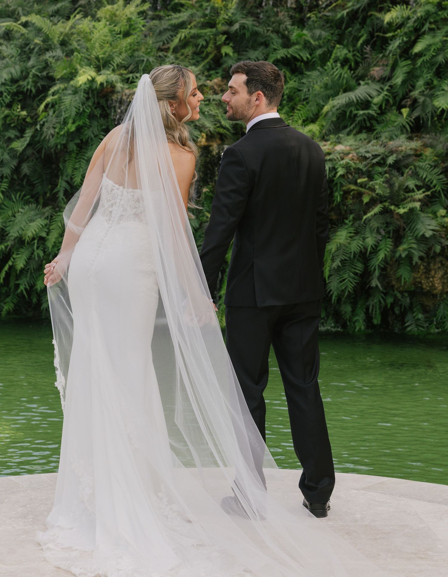 A couple in wedding attire walks together along a scenic waterfront path surrounded by lush greenery.