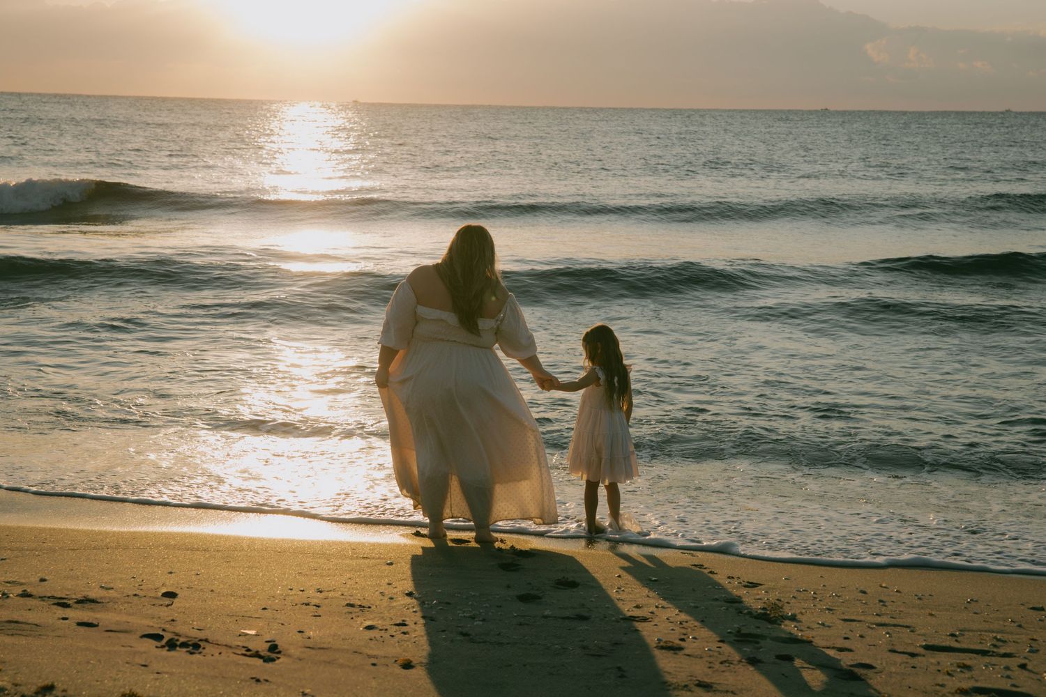 Silhouetted figures walking on beach at sunset with sun glowing over ocean horizon.