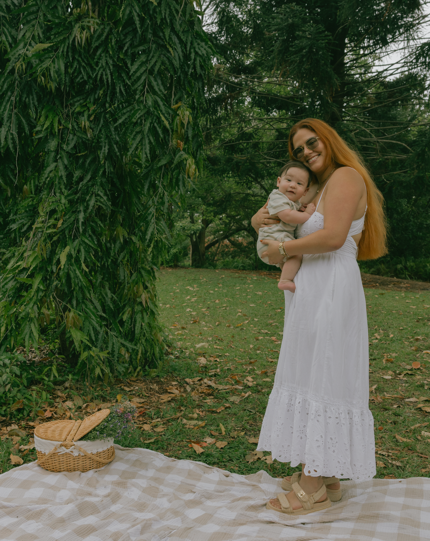 Woman in white dress holding baby while standing on picnic blanket under willow tree.