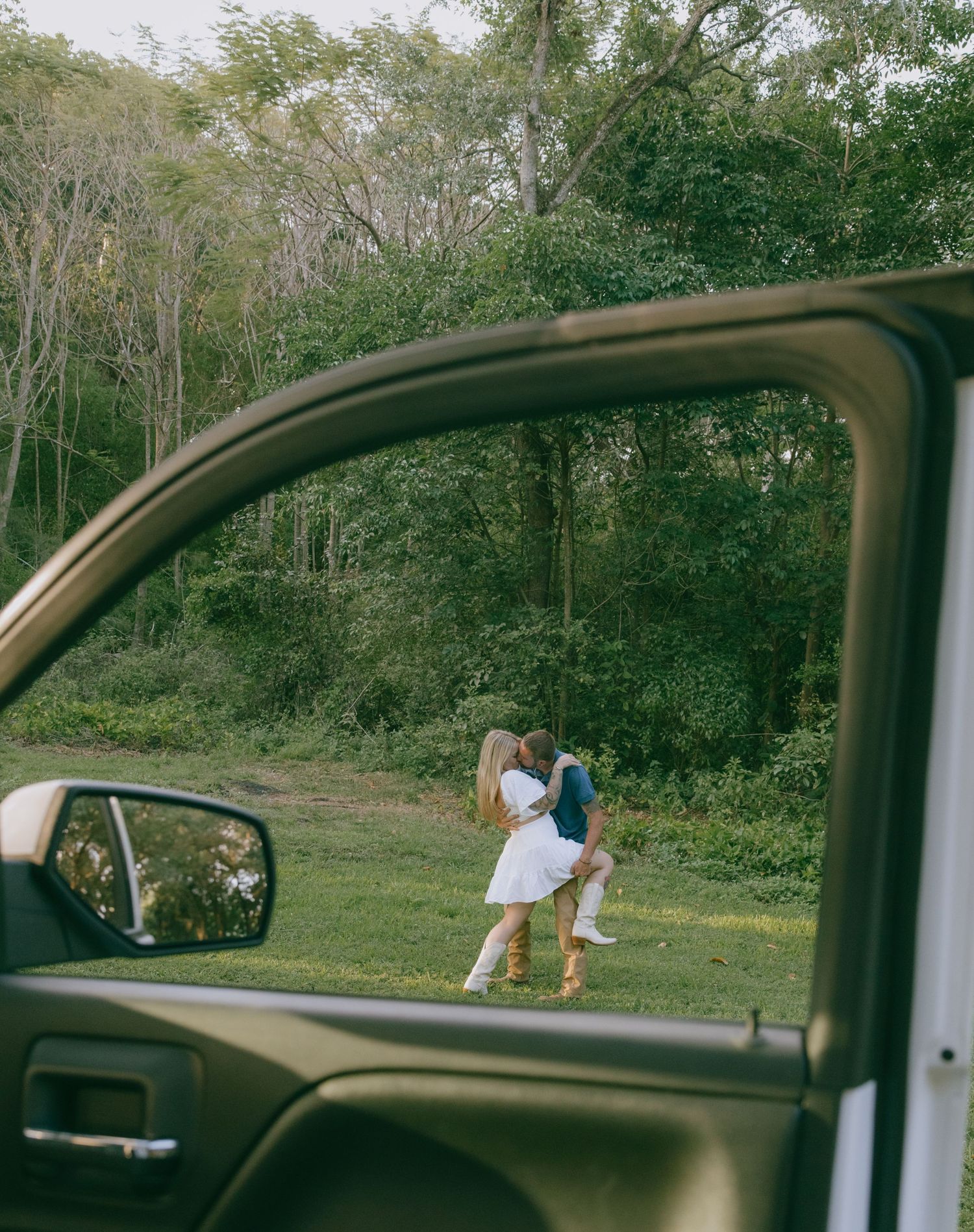 A view through a car window shows a romantic moment in a grassy field surrounded by trees.