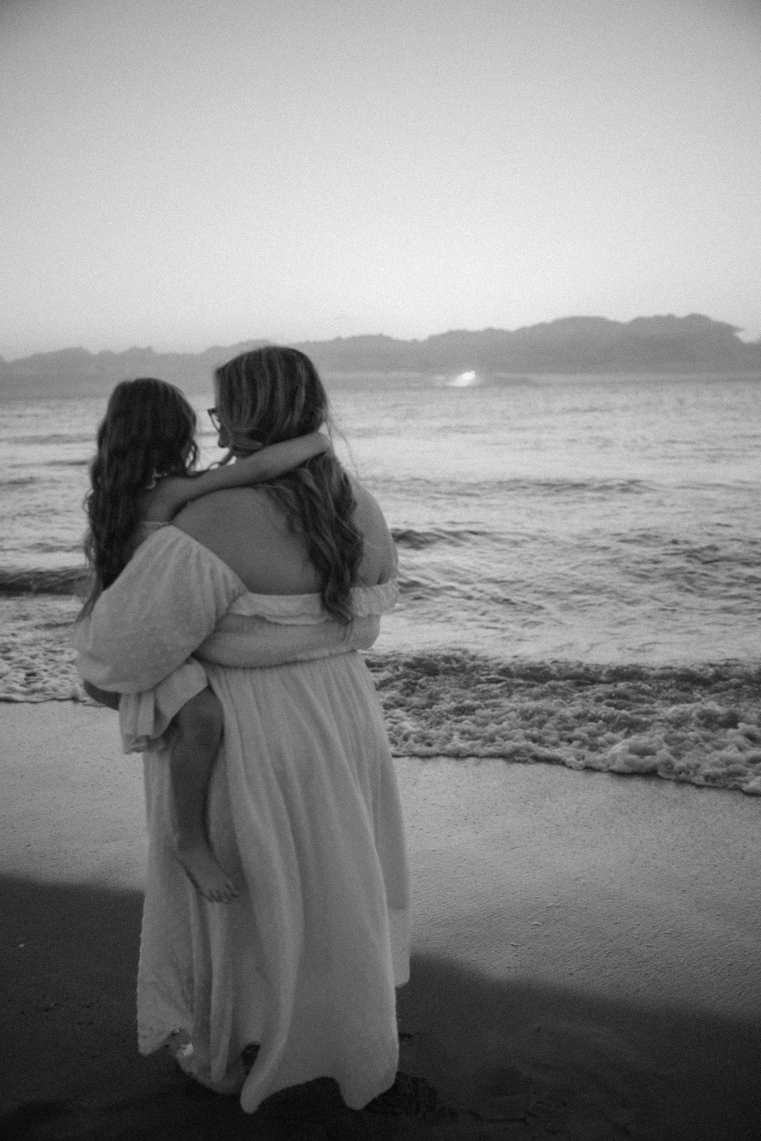 A black and white photo of two people in white dresses sharing an intimate moment by the ocean.