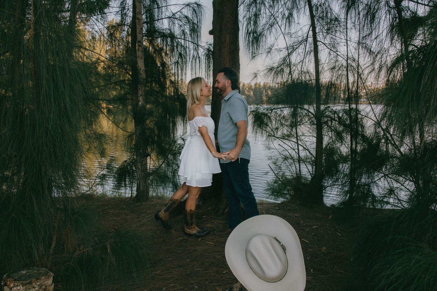 Couple in casual attire walking along wooded lakeside path at sunset.