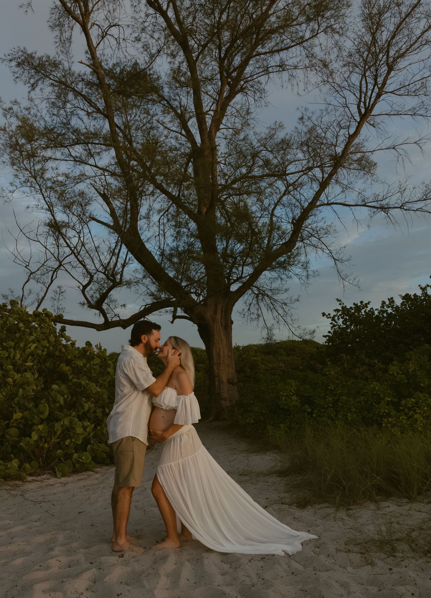 A romantic couple embraces on a sandy beach at sunset with a large tree silhouetted against the moody sky.