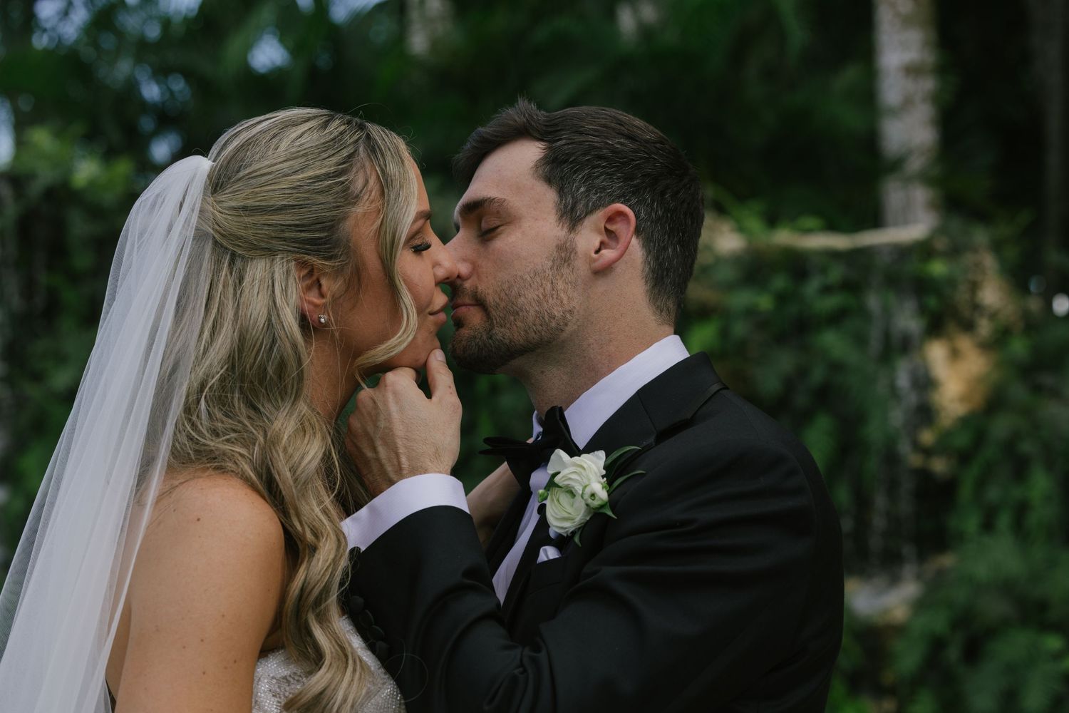 A romantic wedding couple shares a tender kiss in a lush garden setting with natural greenery in the background.